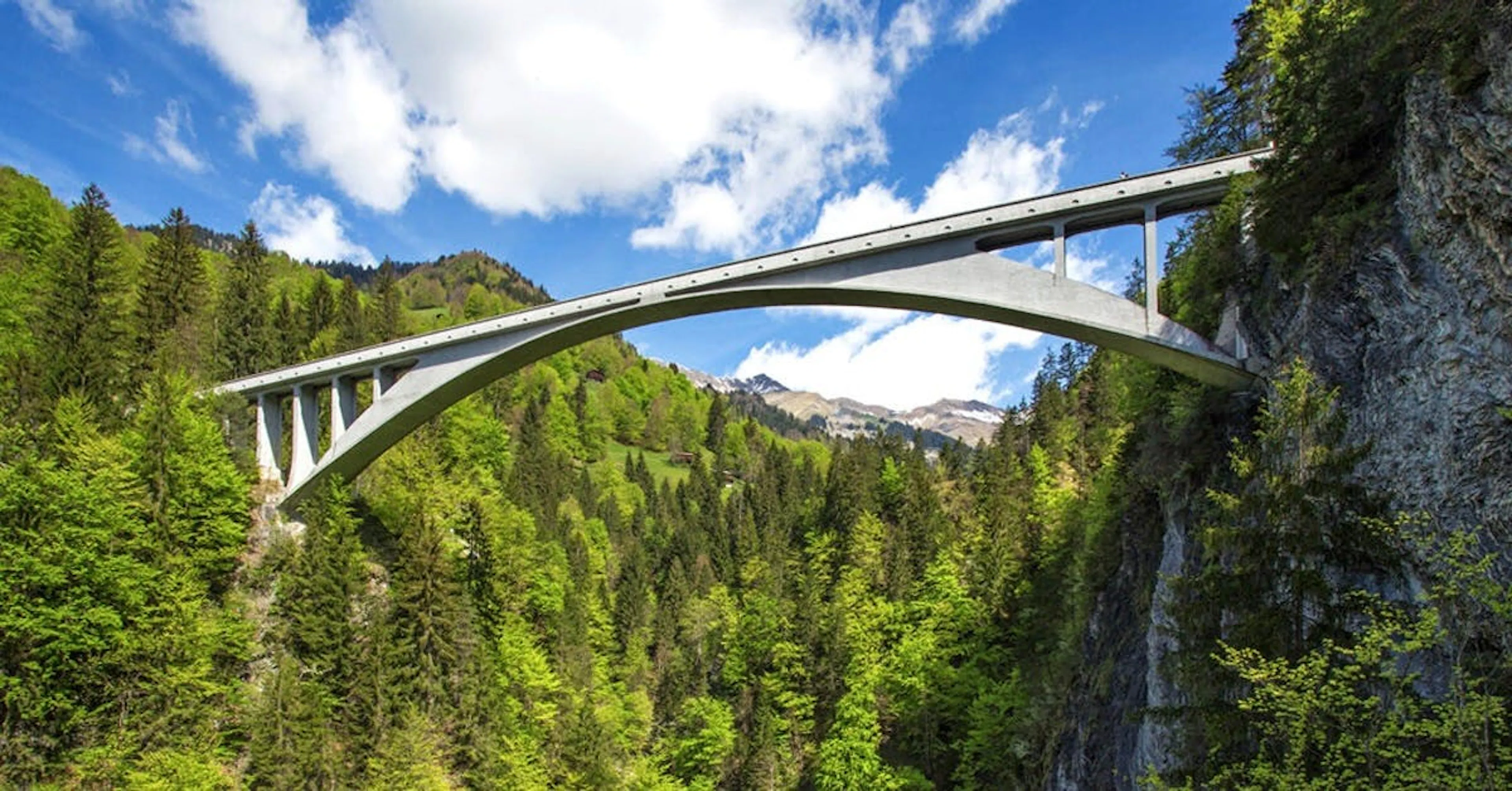 Photograph of a slender white concrete arch bridge spanning a forested alpine gorge
