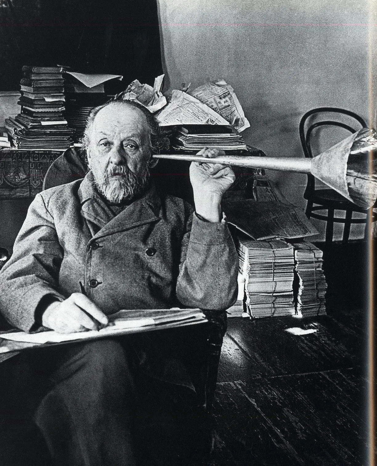 Photograph of Konstantin Tsiolkovsky at his paper-covered desk holding a model rocket nozzle