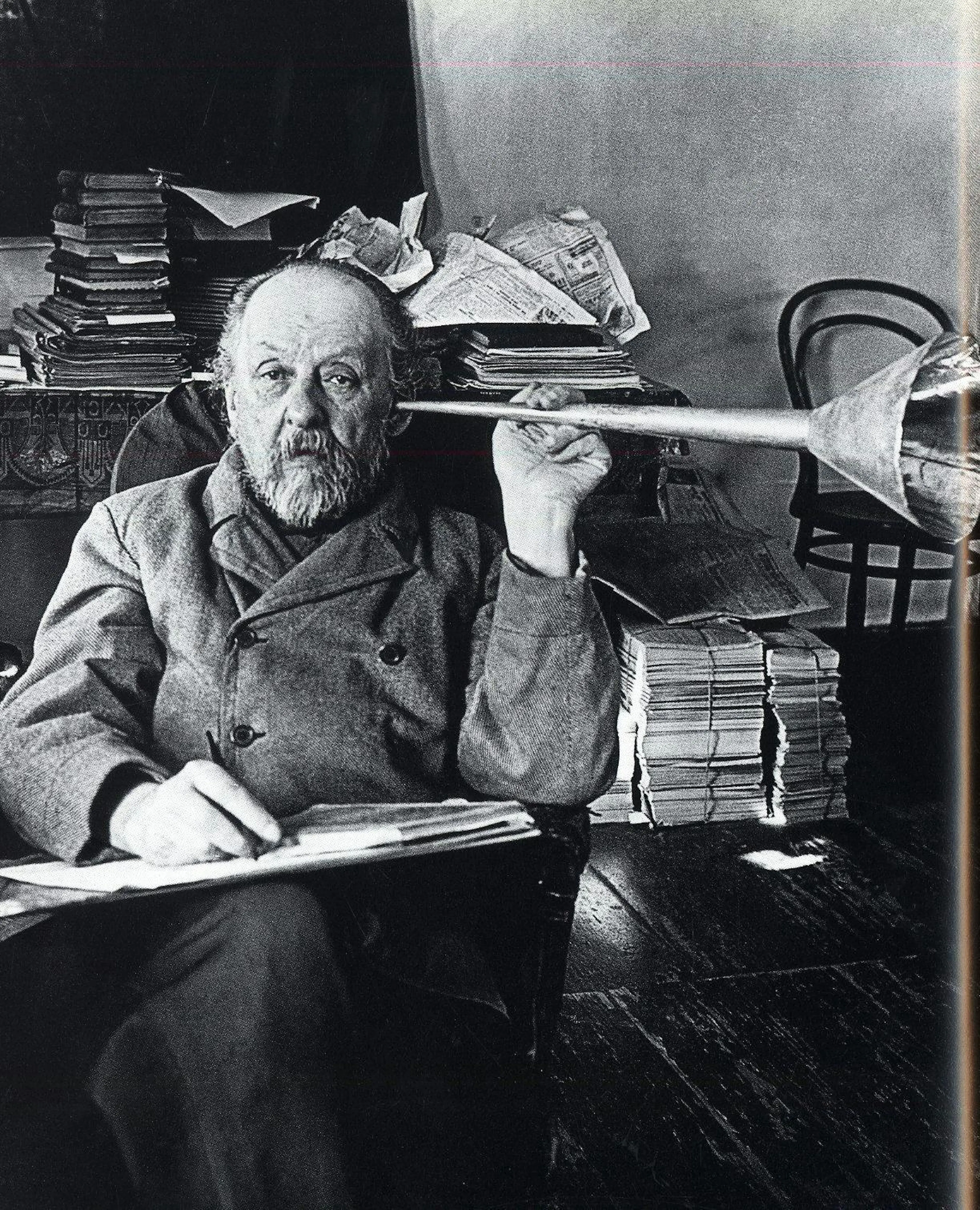 Photograph of Konstantin Tsiolkovsky at his paper-covered desk holding a model rocket nozzle