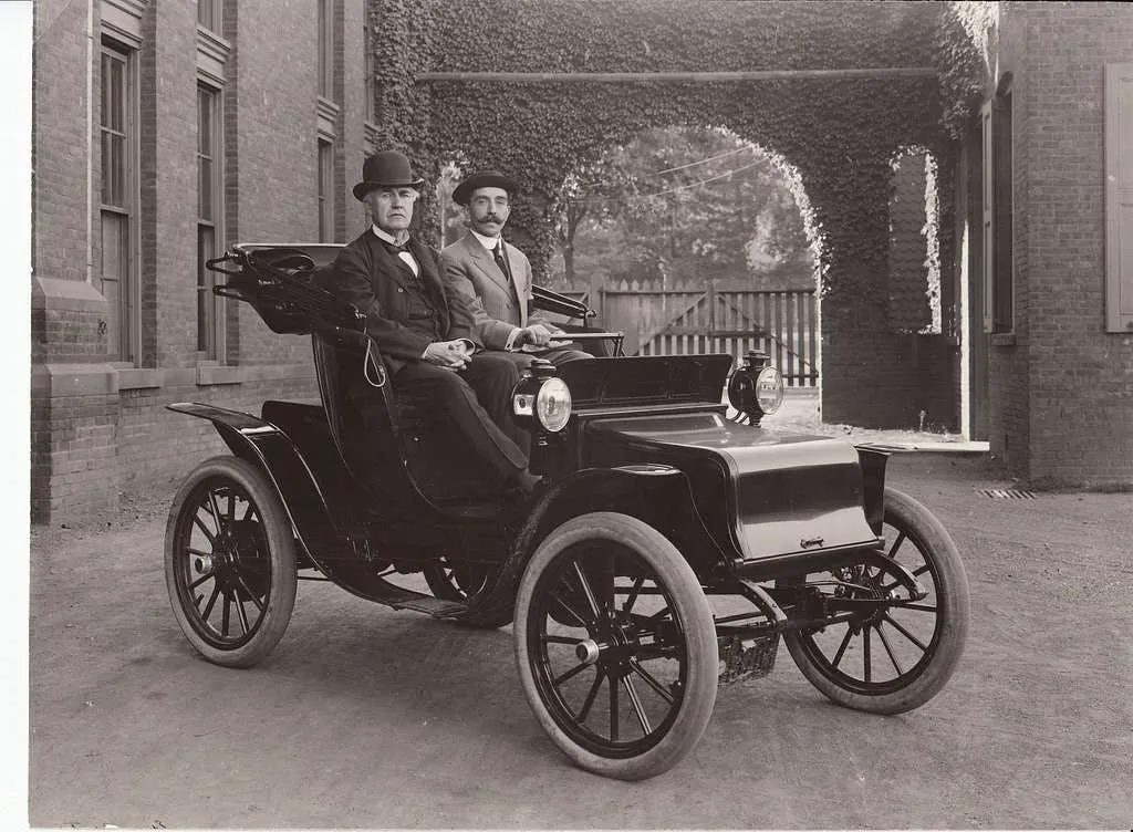 Black-and-white photograph of two men in bowler hats seated in an early electric car outside a brick building