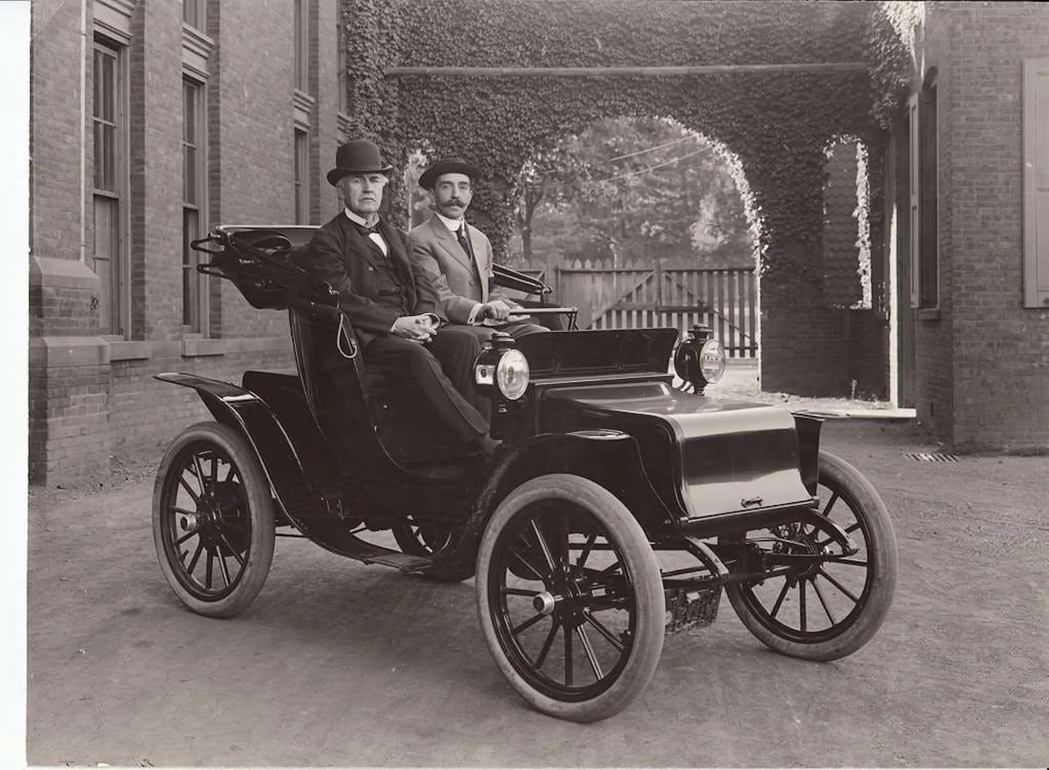 Black-and-white photograph of two men in bowler hats seated in an early electric car outside a brick building
