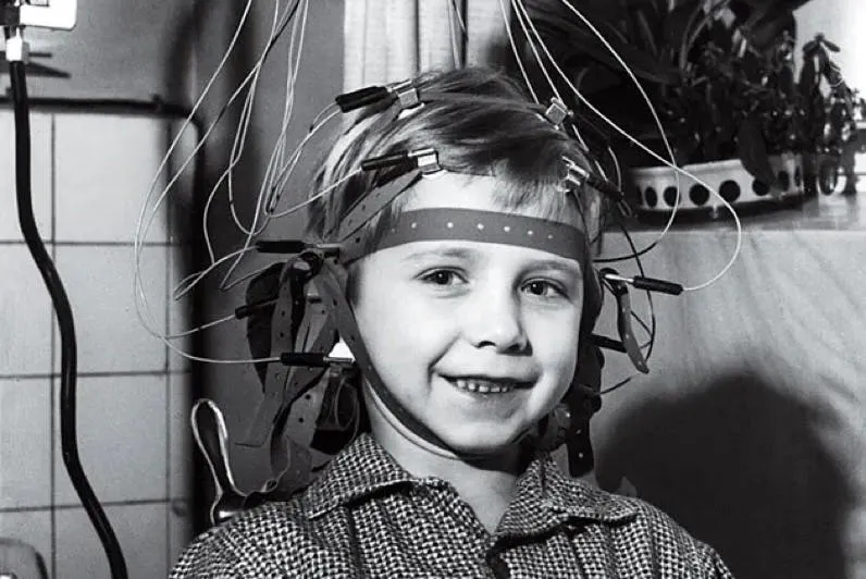Black-and-white photograph of a smiling boy wearing an early EEG headset trailing electrode wires