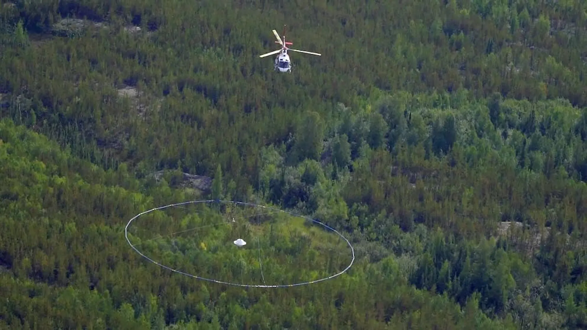 Helicopter towing a large circular transmitter loop over forest during an airborne geophysical survey
