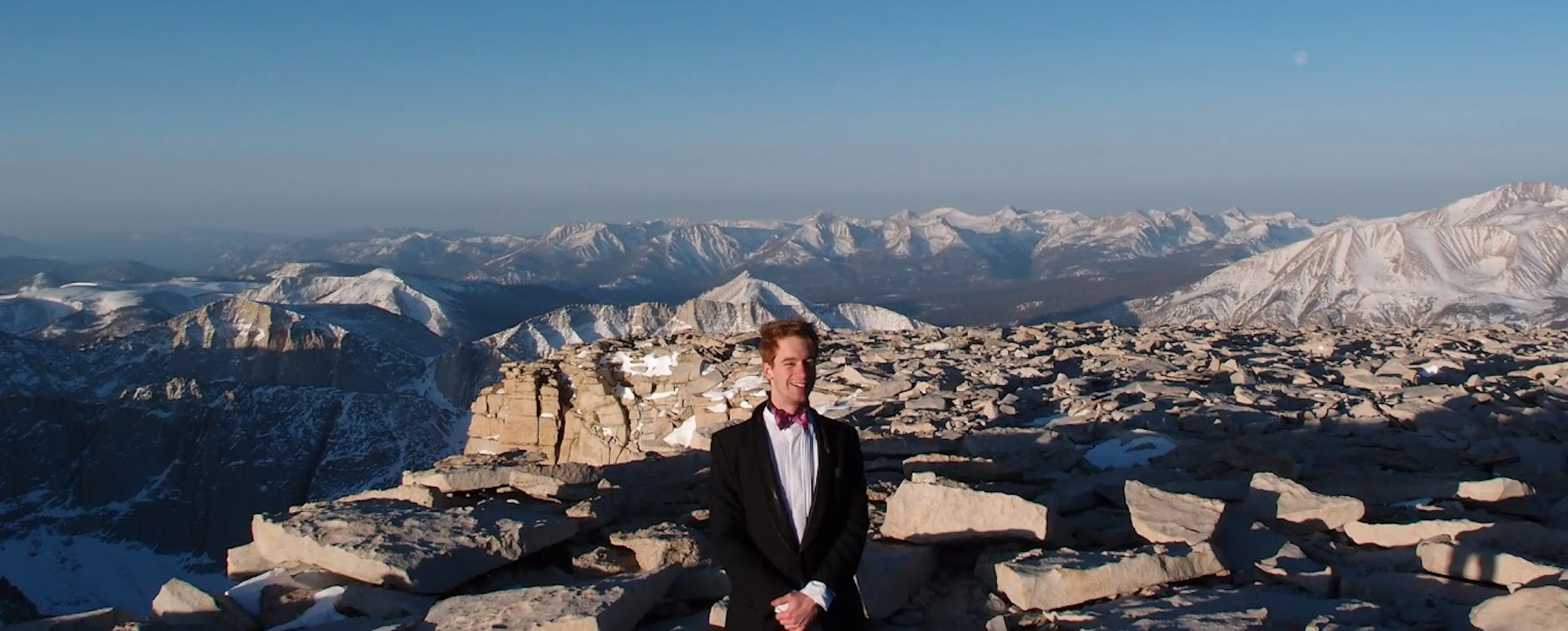 Casey Handmer, founder of Terraform Industries, standing in a suit and bow tie on a rocky, snow-dusted mountain summit