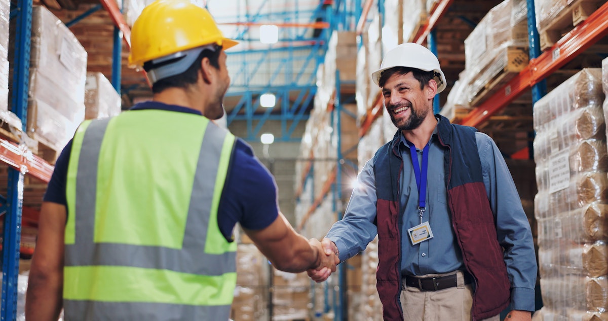 Two men in reflective vests shaking hands in a warehouse