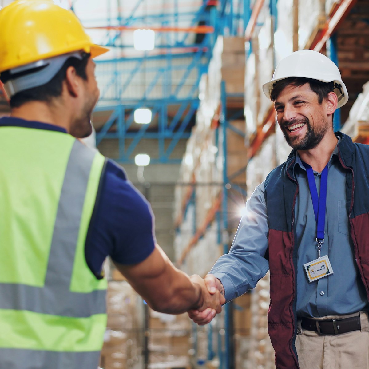 Two men in reflective vests shaking hands in a warehouse