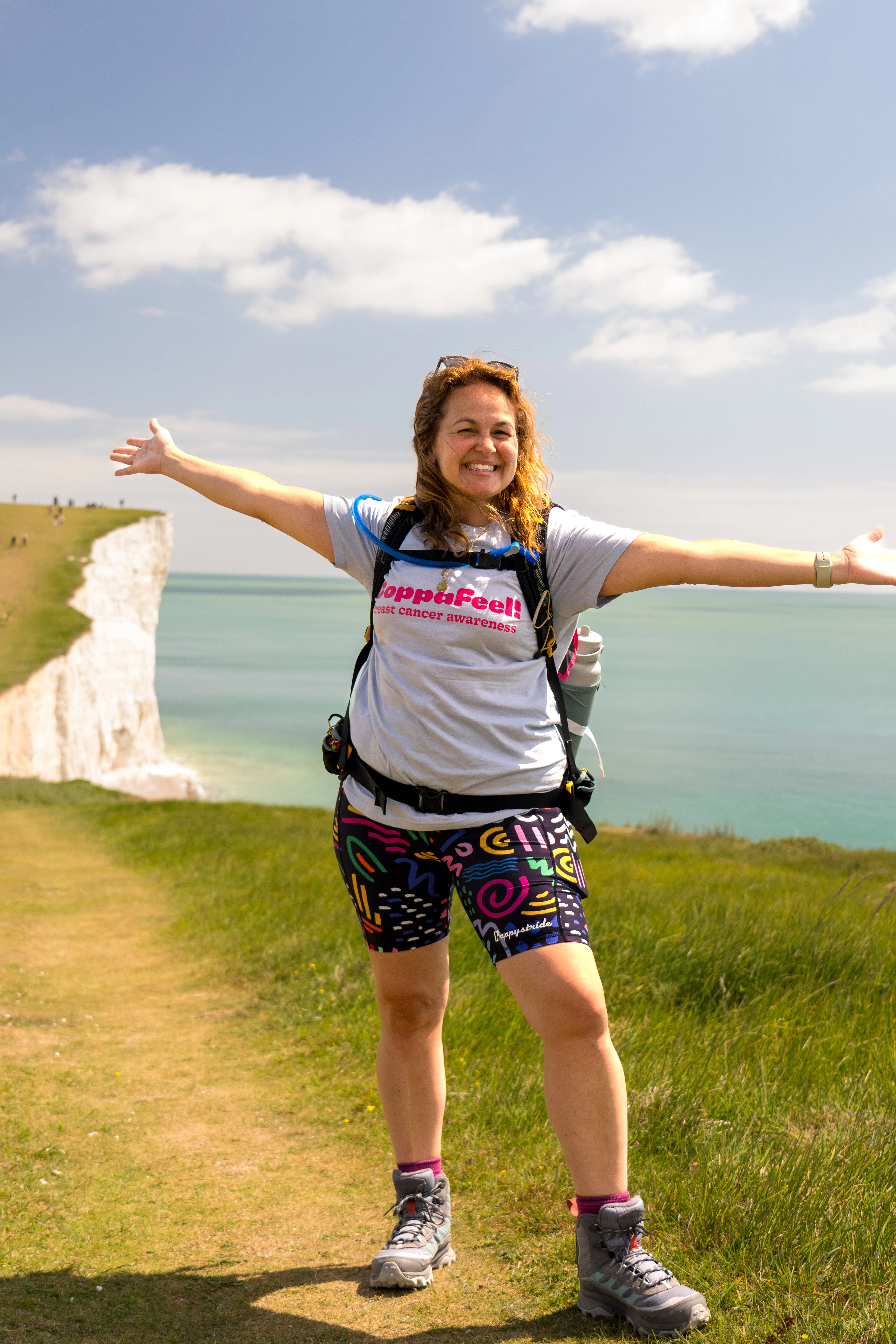Gi smiling, with arms outstretched, standing in front of the coastline, in a CoppaFeel t-shirt.