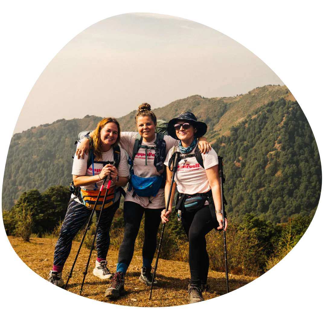 Three hikers standing close together on a grassy mountain trail, wearing matching white T-shirts with printed text, backpacks, and hiking gear. Each person holds trekking poles. Behind them, rolling green hills and forested slopes stretch into the distance under a hazy sky.