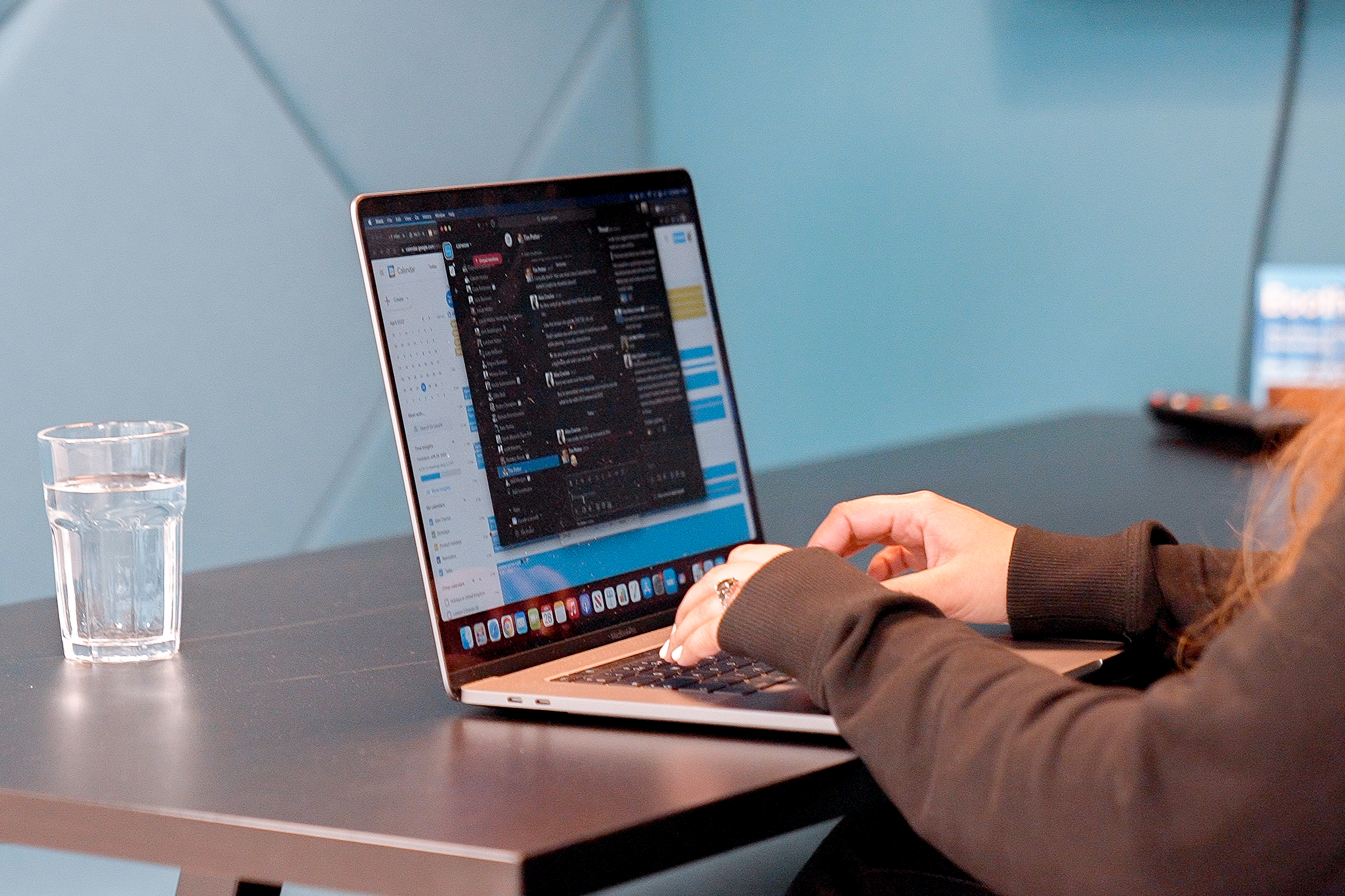 A laptop on a metal desk in a modern office, with the arms of the person working on it visible in the shot