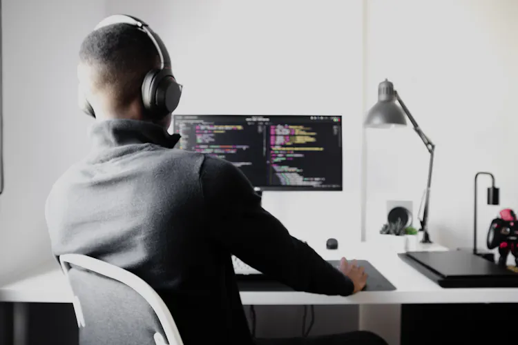 Cover of Man sitting at a desk with code on monitor, wearing headphones article