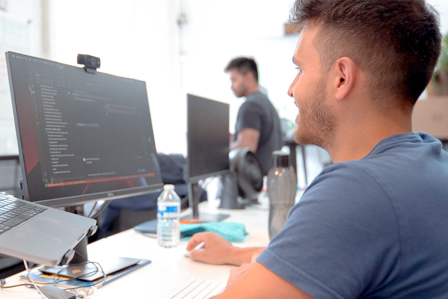 A man seen from behind in a brightly-lit office looking at a monitor with code on it.