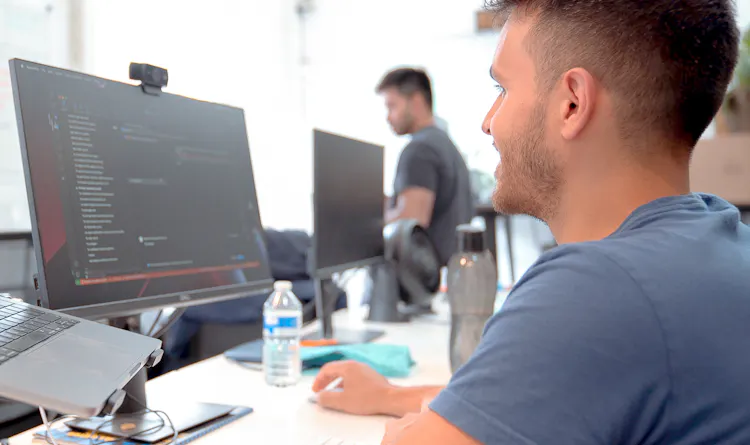 A man seen from behind in a brightly-lit office looking at a monitor with code on it.