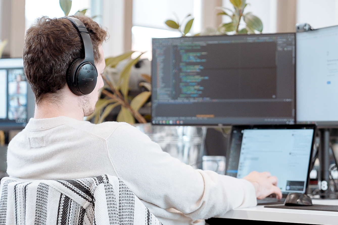 Cover of A man sitting with his back to the camera looking at a laptop and two monitors with code on them article