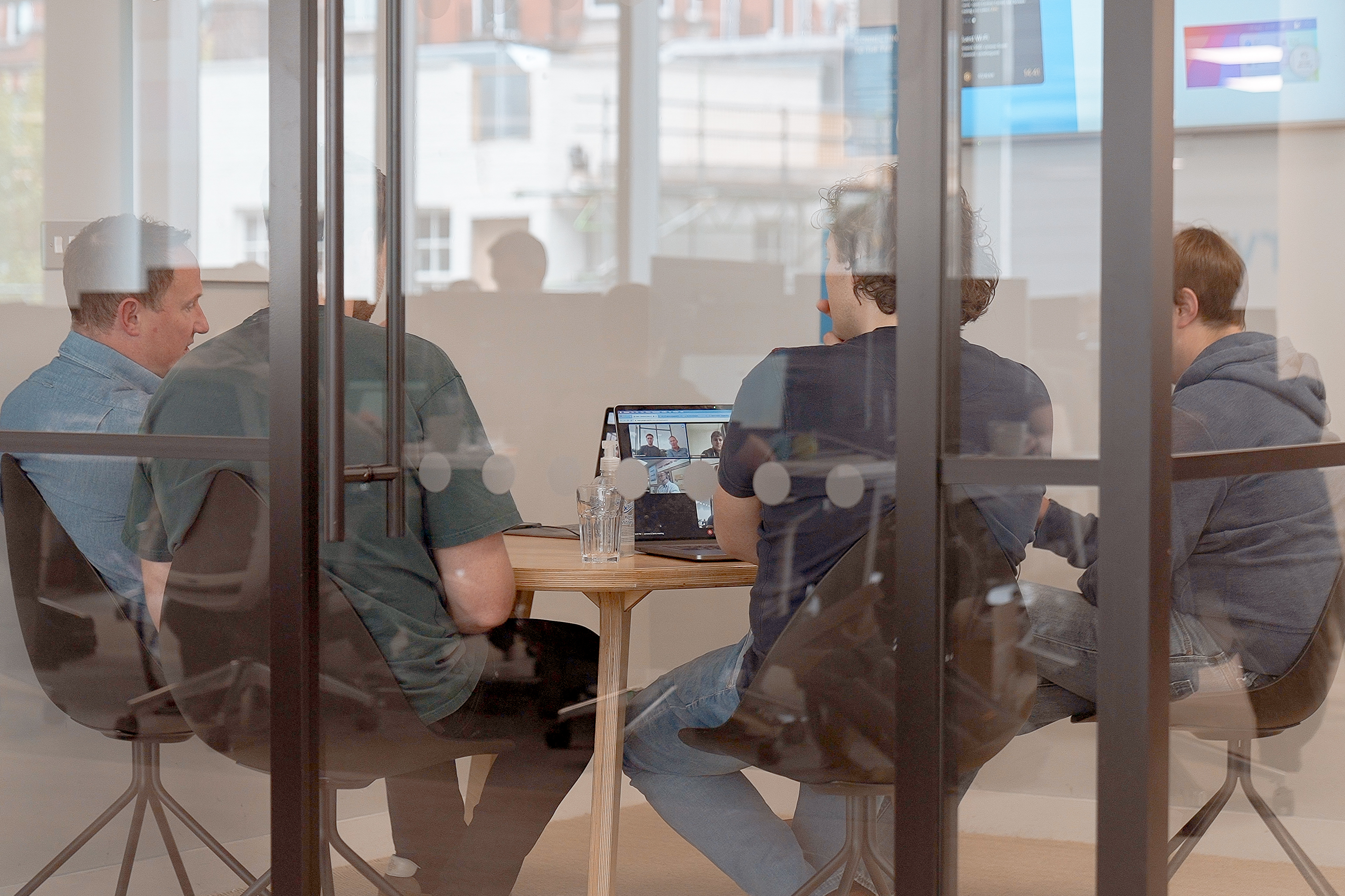 Four men sitting around a table on a conference call are visible through a glass partition. Their backs are turned to the camera and the image is partially obscured by reflections on the window