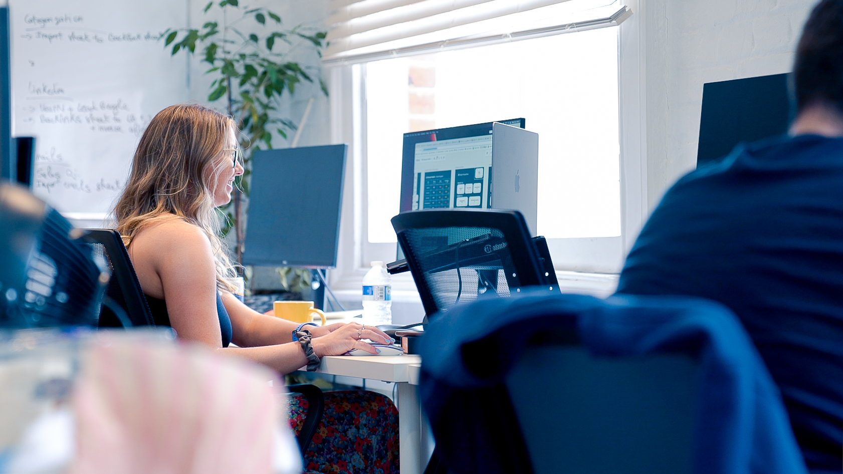 Cover of A woman smiles while working at a laptop on a bank of desks in an office article