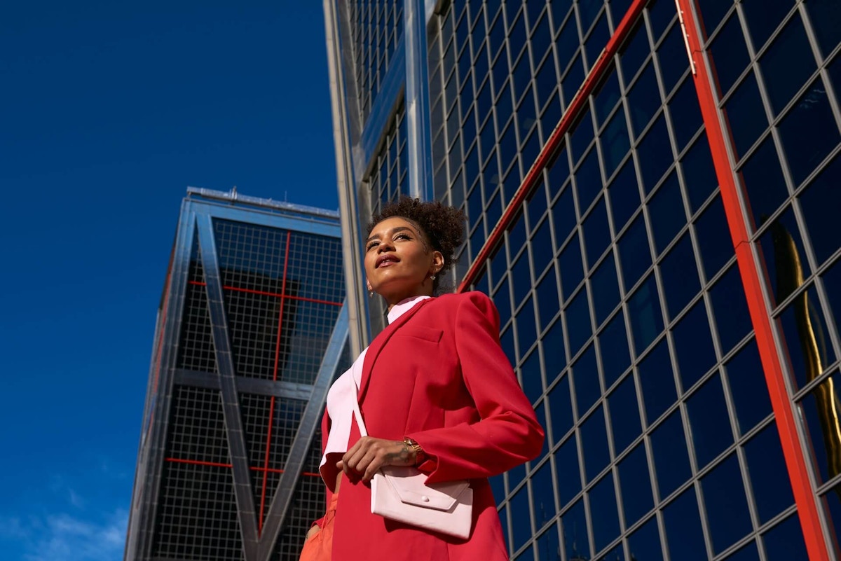 Lady in a red business suit and pink purse stands near a glass building, probably a skyscraper