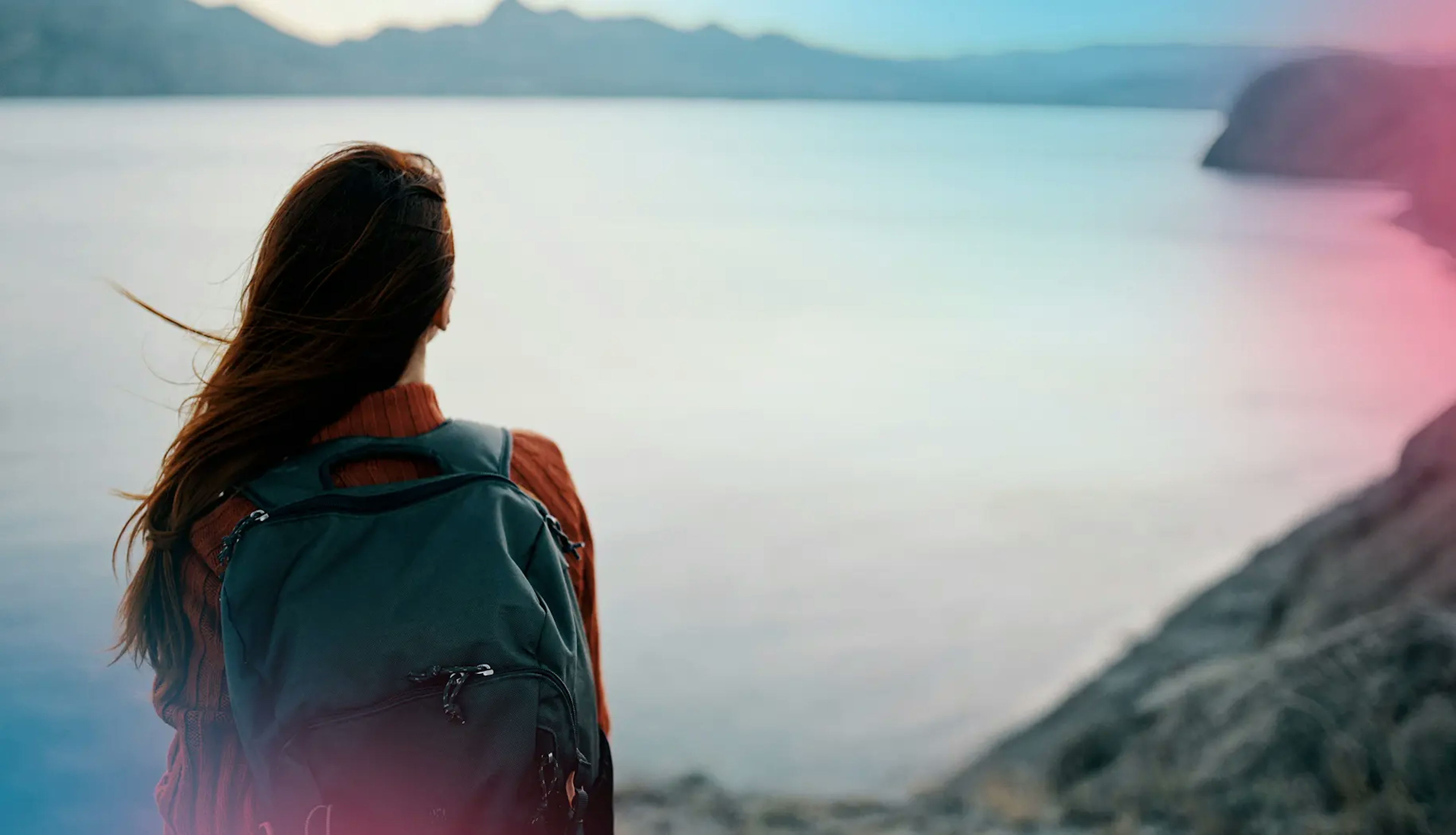 Person with backpack looking out over a wide lake, symbolizing focus, vision, and forward-looking determination to create real-world impact.