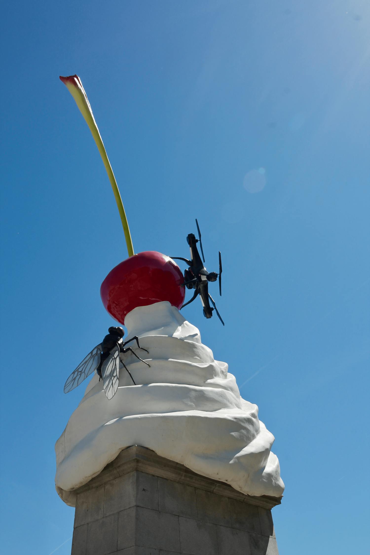 [ID: This photograph shows a sculpture in the form of a pile of white cream topped with a red cherry with a tall green stem. A black fly clings onto the cream while a black drone is perched on the side of the cherry. The sculpture is placed on top of a rectangular grey stone plinth against a background of a blue, cloudless sky. End of ID]