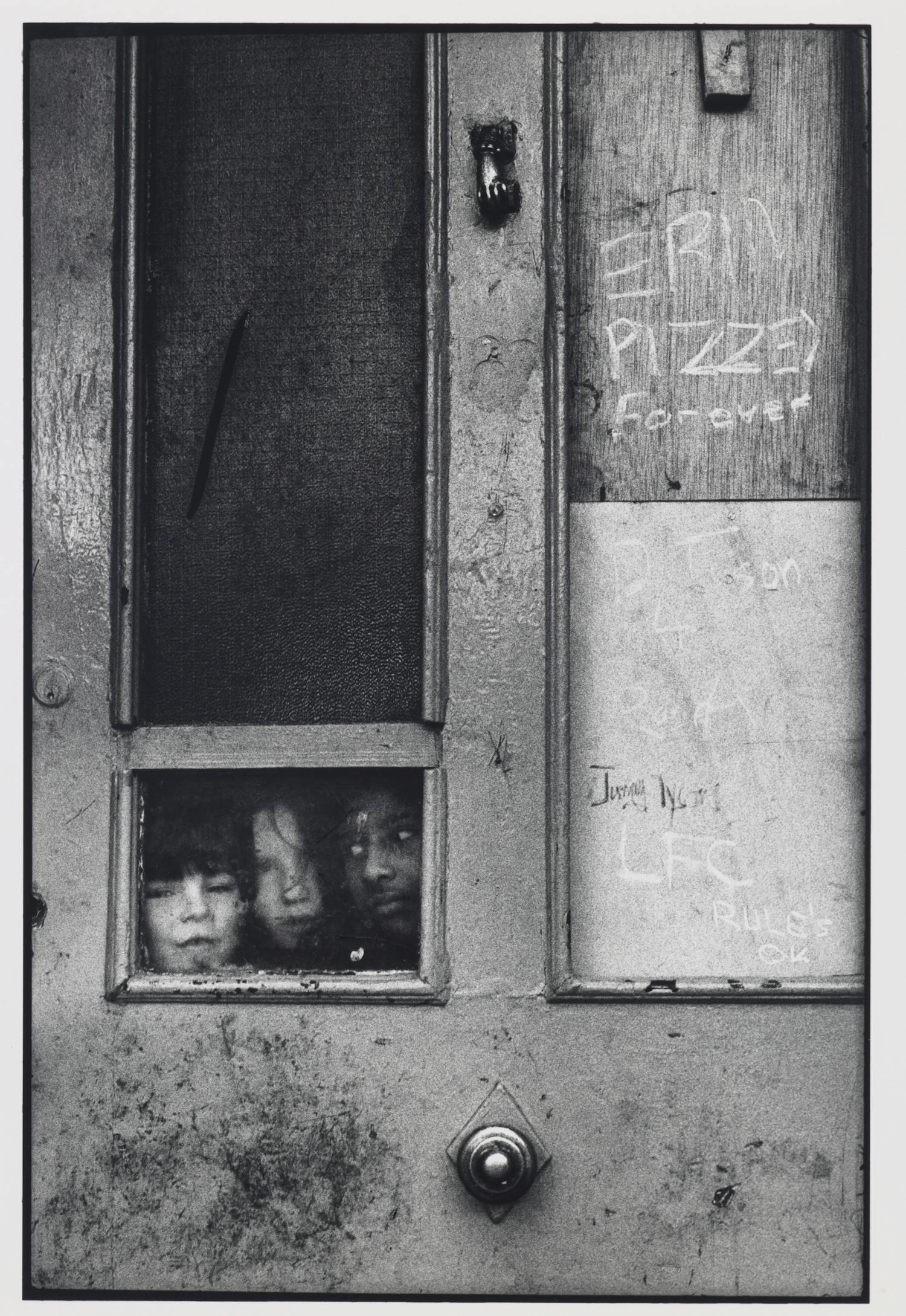 [ID: The black and white photograph shows an old door covered with graffiti. Three children are looking through a small window embedded in the door. End of ID]