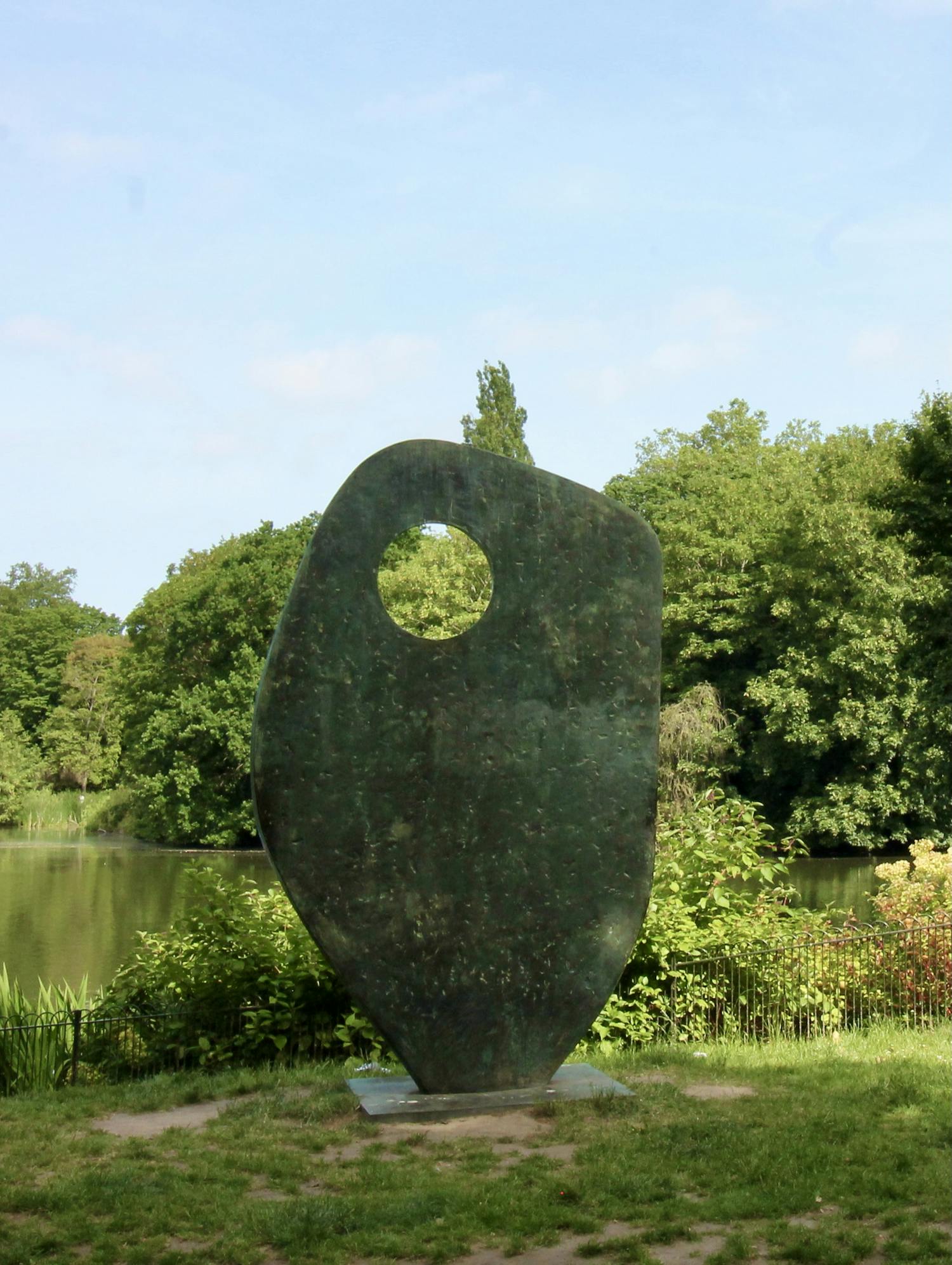 [ID: This photograph shows an abstract brass sculpture in an angular ovoid form with a circular hole in the top left-hand corner. The sculpture is wider at the top and tapers at the bottom where it meets the concrete base it is placed on. The sculpture is located on a grassy bank next to a lake beyond which a line of trees can be seen. End of ID]