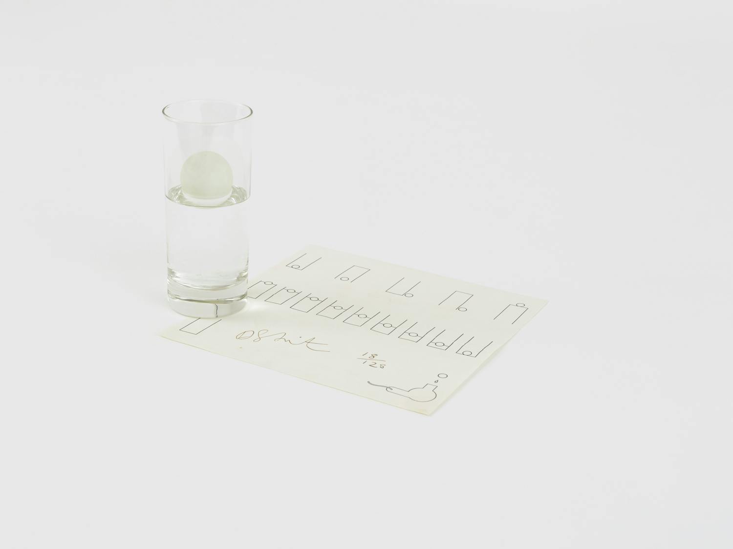 [ID: The image shows a transparent glass half filled with water; a ping pong ball is floating on the surface. The glass sits on top of a white sheet of paper. On the paper, the artist has drawn a series of diagrams that represent the possible ways for the glass, ping pong ball and water to interact. End of ID]