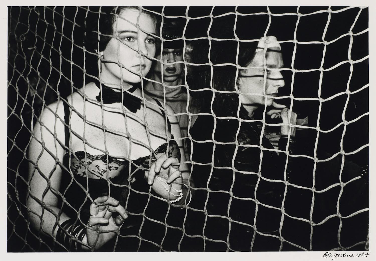 [ID: The black and white photograph shows a person standing in the foreground behind a net. They are wearing a bow-tie and a corset, and their hands are gripping the net. Behind them there are two other people, their faces barely visible as they emerge from the dark background. End of ID]