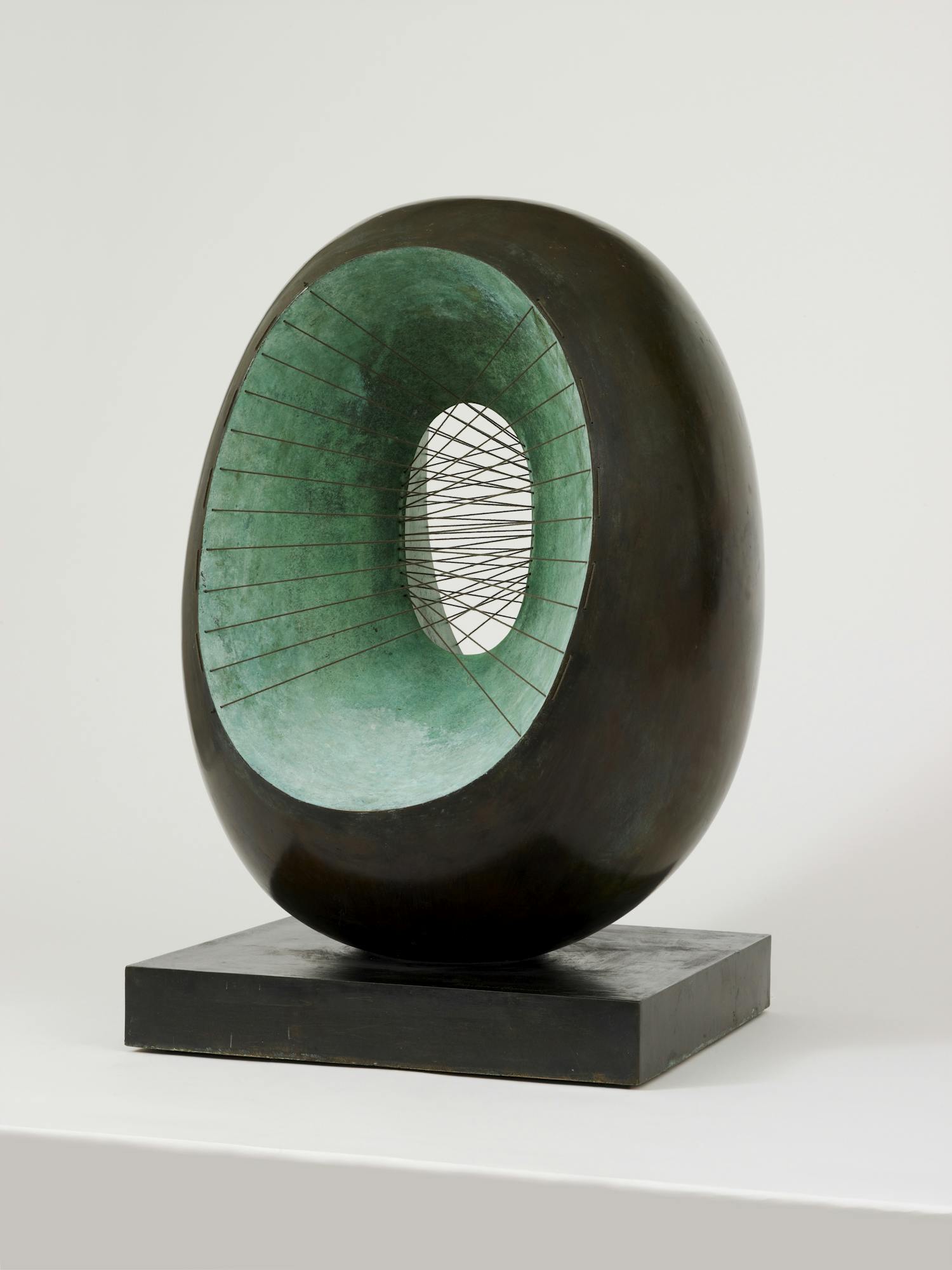 [ID: The image shows an oval shaped black sculpture with an oval opening, revealing its green interior. A series of strings are stretched from left to right and through the sculpture. End of ID]