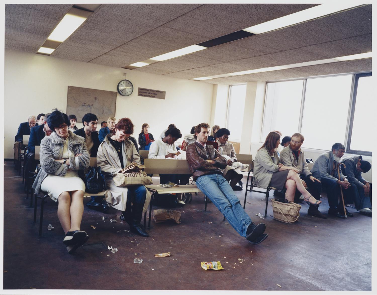 [ID: The image shows a barren room with a window front on the right side. People are sitting on benches which are placed throughout the room. End of ID]