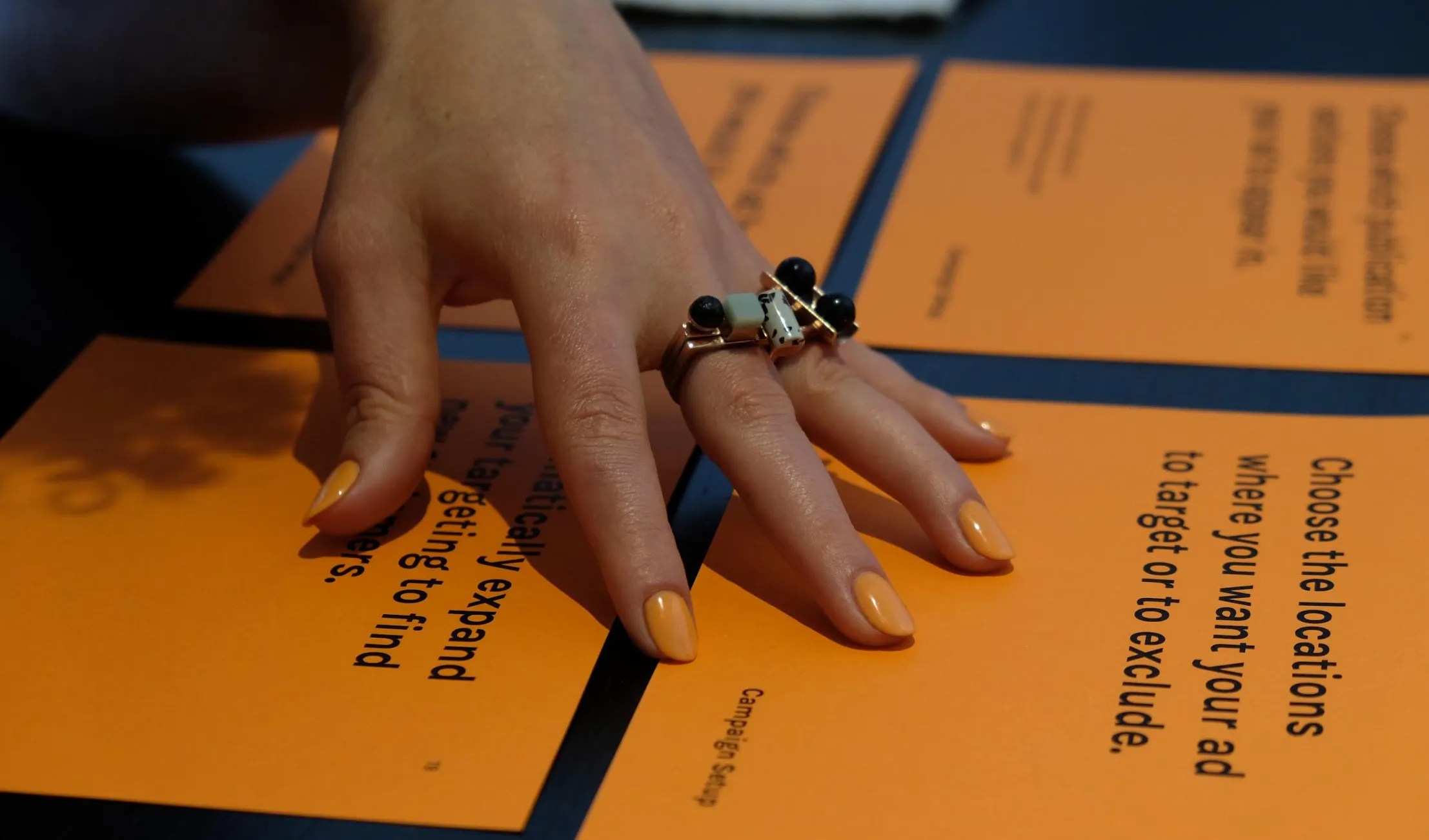 A hand with orange-painted nails and a distinctive geometric ring rests on bright orange cards for Strategic research and prototyping for a Google-powered Fairfax Media self-serve SME advertising platform, a project by CraigWalker (CW) a future service studio. The cards display bold black text with advertising-related prompts, such as “Choose the locations where you want your ad to target or to exclude.