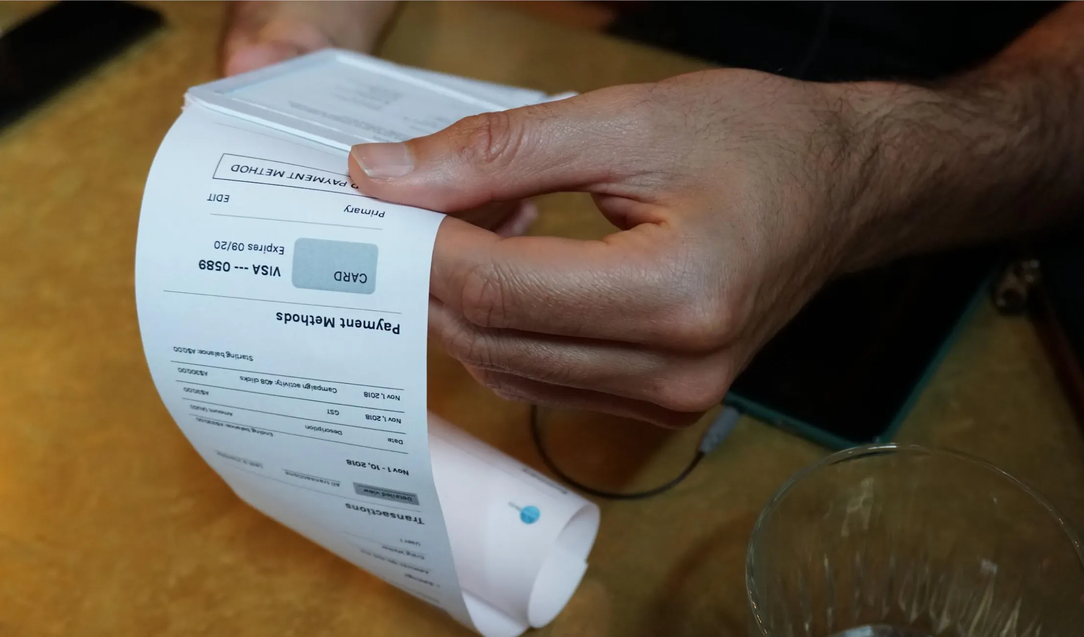 A close-up of a person’s hand holding a printed payment receipt that shows transaction details and payment methods. A glass of water and part of an electronic device are visible on the table in the background. For Strategic research and prototyping for a Google-powered Fairfax Media self-serve SME advertising platform, a project by CraigWalker (CW) a future service studio.