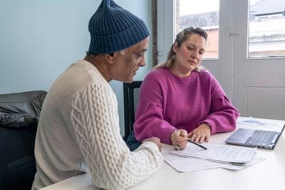 A man and woman sit together looking through paperwork. The woman is holding a pen and gesturing to the text on the paper, as if explaining.