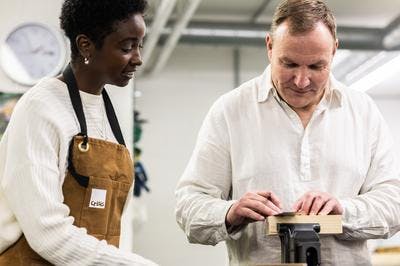 A man and woman stand together near a tool bench. The man holds a pencil and is looking at his work, while the woman looks on wearing a Crisis apron.