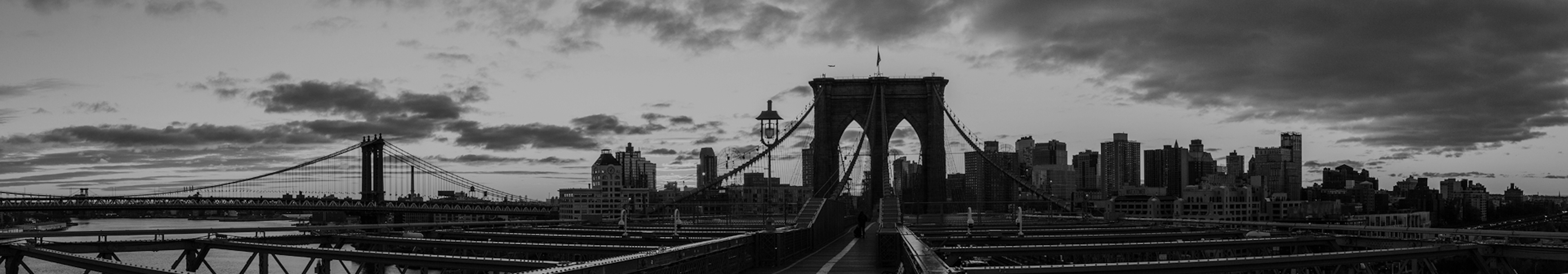 Black and white photo of the Brooklyn Bridge walkway at dawn or dusk, with dramatic clouds overhead and the Manhattan skyline in the background. The bridge’s iconic stone arches and suspension cables create strong leading lines toward the horizon.