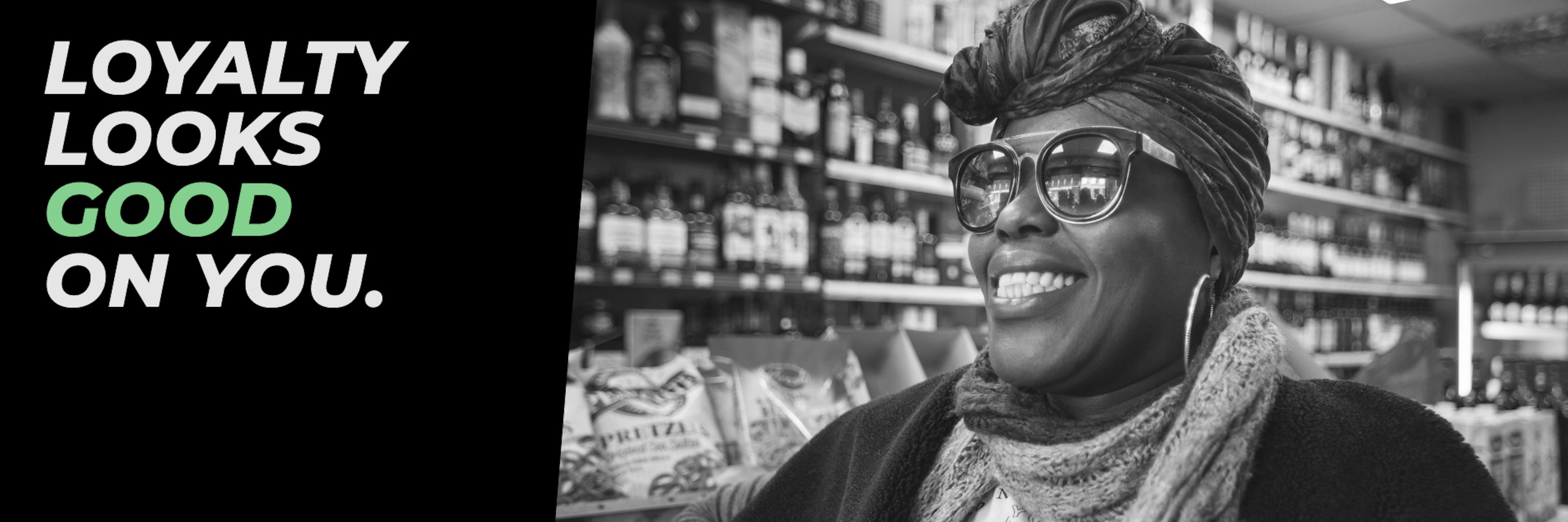 Black and white photo of a smiling woman wearing sunglasses and a headwrap, standing in a liquor store aisle with shelves of bottles behind her. Bold text on the left reads “LOYALTY LOOKS GOOD ON YOU,” with the word “GOOD” highlighted in green.