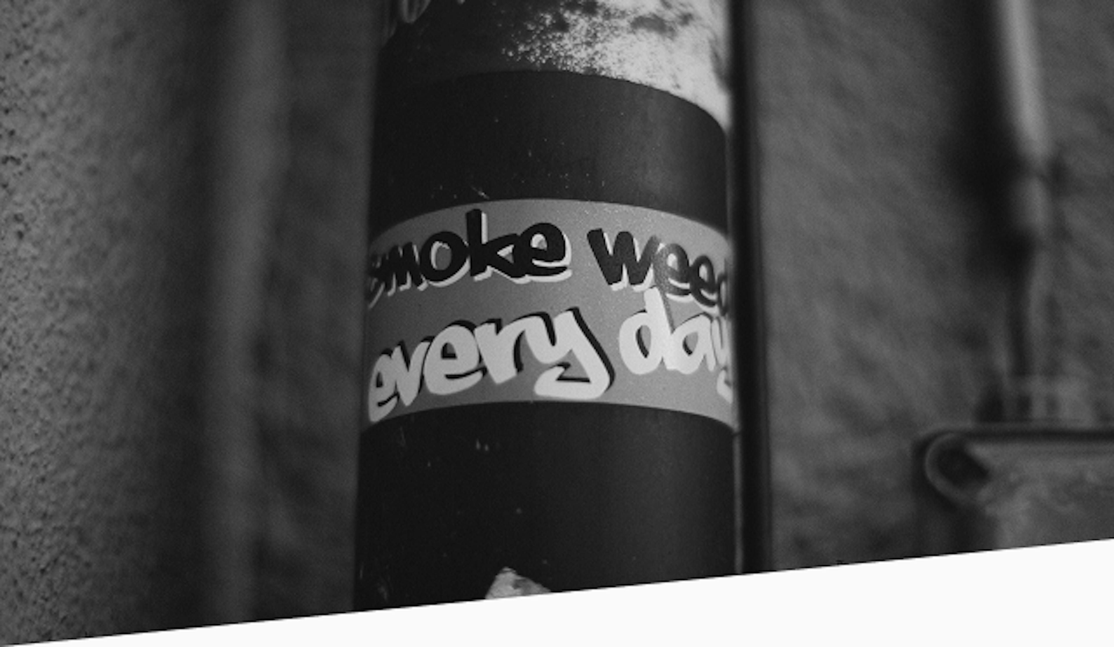 Black and white photo of a street pole with a graffiti-style sticker that reads “smoke weed every day,” placed against a textured wall with visible urban wear.