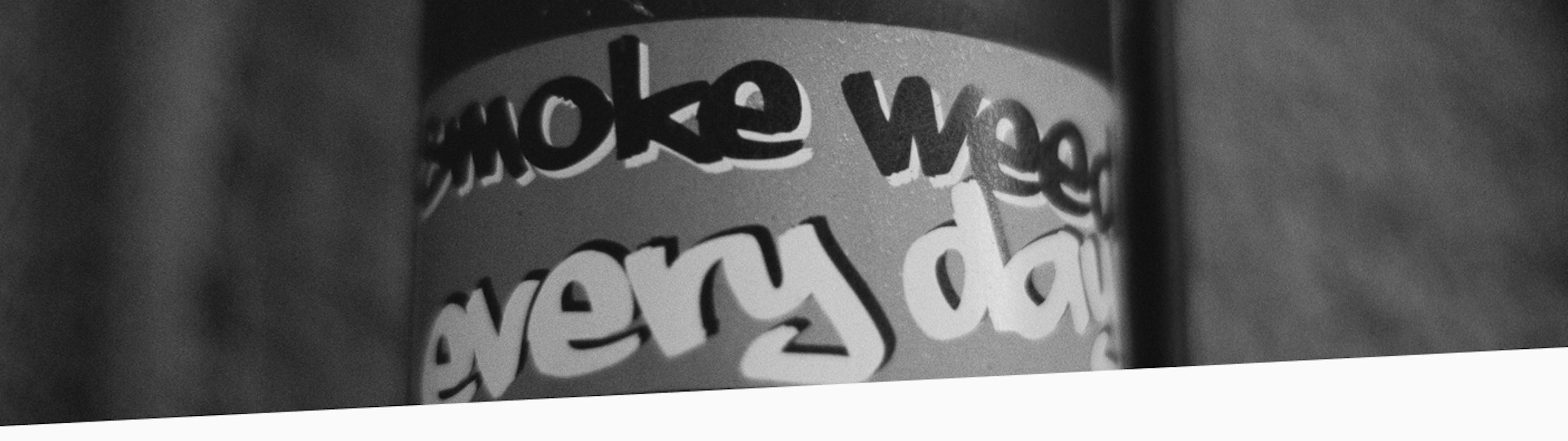 Black and white photo of a street pole with a graffiti-style sticker that reads “smoke weed every day,” placed against a textured wall with visible urban wear.