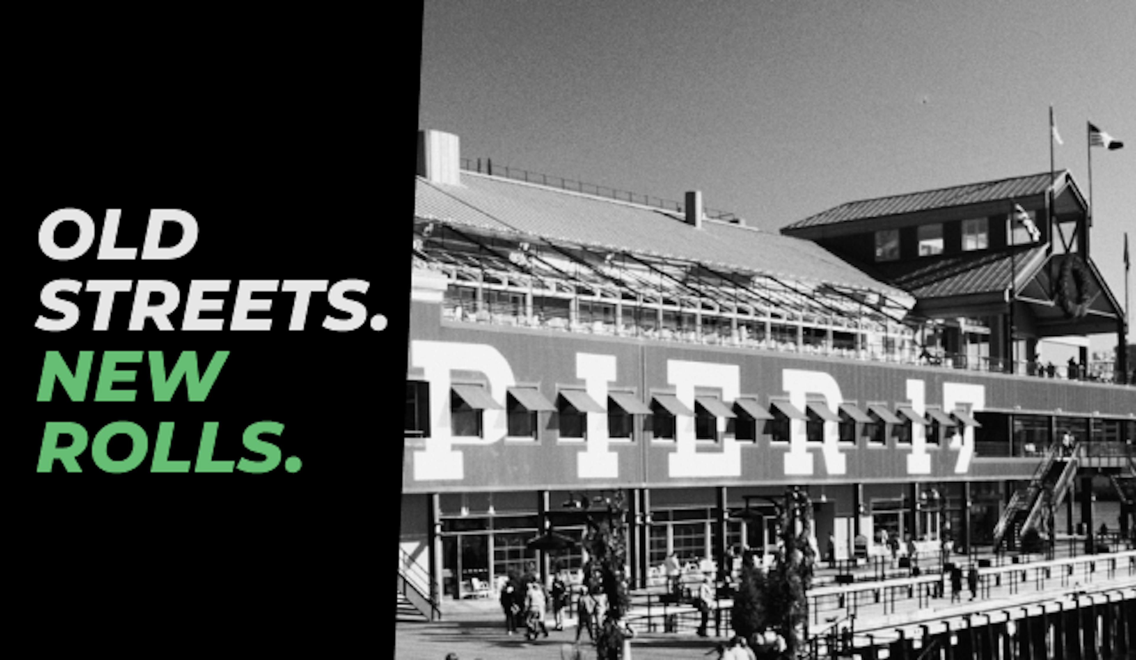 Black and white photo of Pier 17 at the South Street Seaport in New York City, with crowds walking along the boardwalk and a docked boat in the water. Bold text on the left reads: “OLD STREETS. NEW ROLLS.” with “NEW ROLLS.” highlighted in green.
