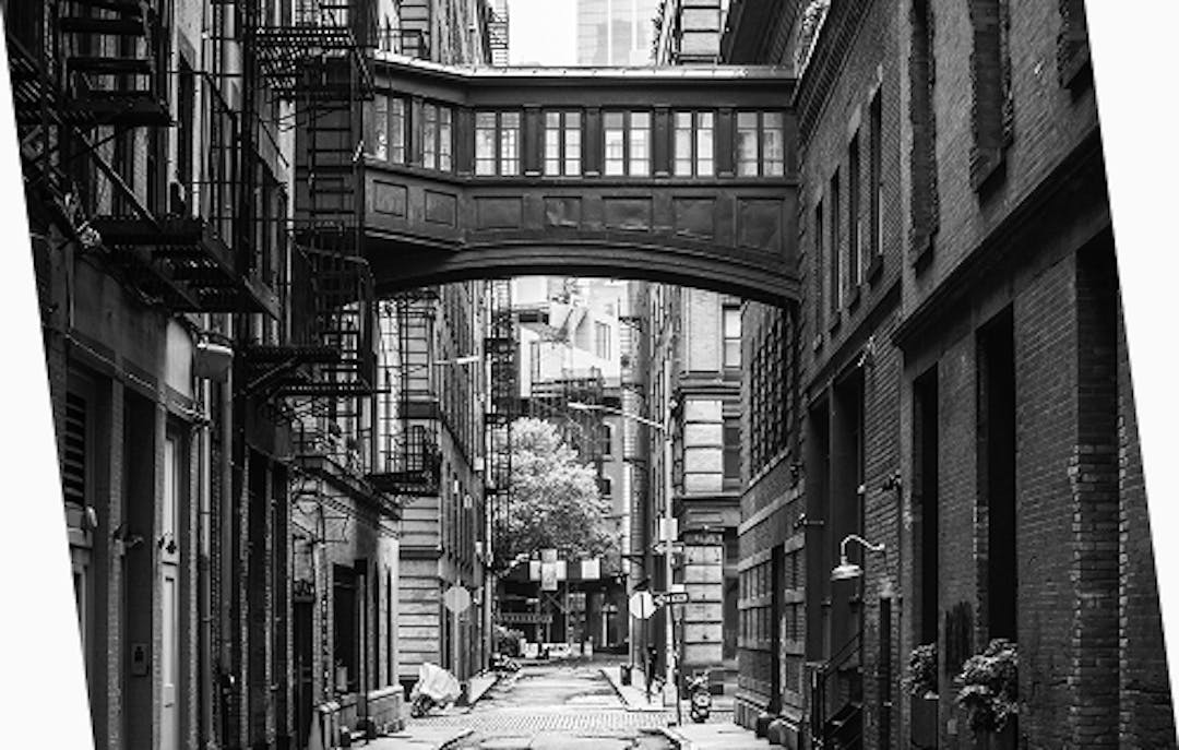 Daytime alley scene in Tribeca, New York, with the iconic elevated pedestrian bridge connecting buildings, surrounded by classic brick facades and fire escapes.