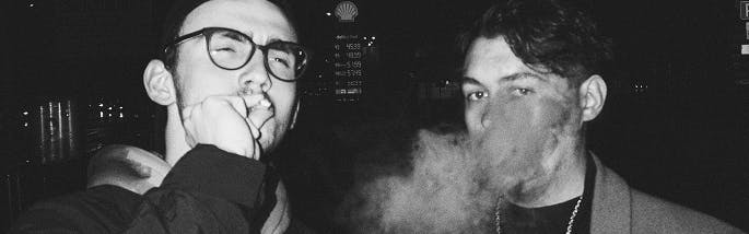 Black and white photo of two young men smoking at night, one exhaling vapor while the other holds a joint to his mouth; urban backdrop with dim signage and streetlights.