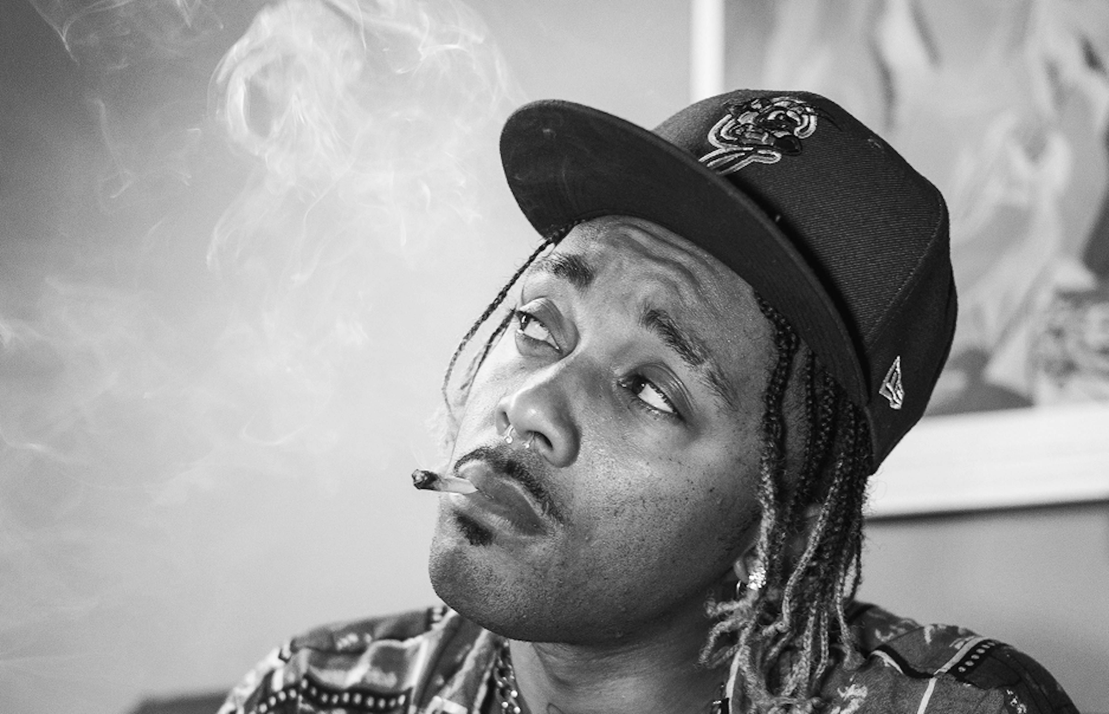 Black and white portrait of a man smoking a joint, wearing a patterned shirt and baseball cap, with smoke swirling around his face.
