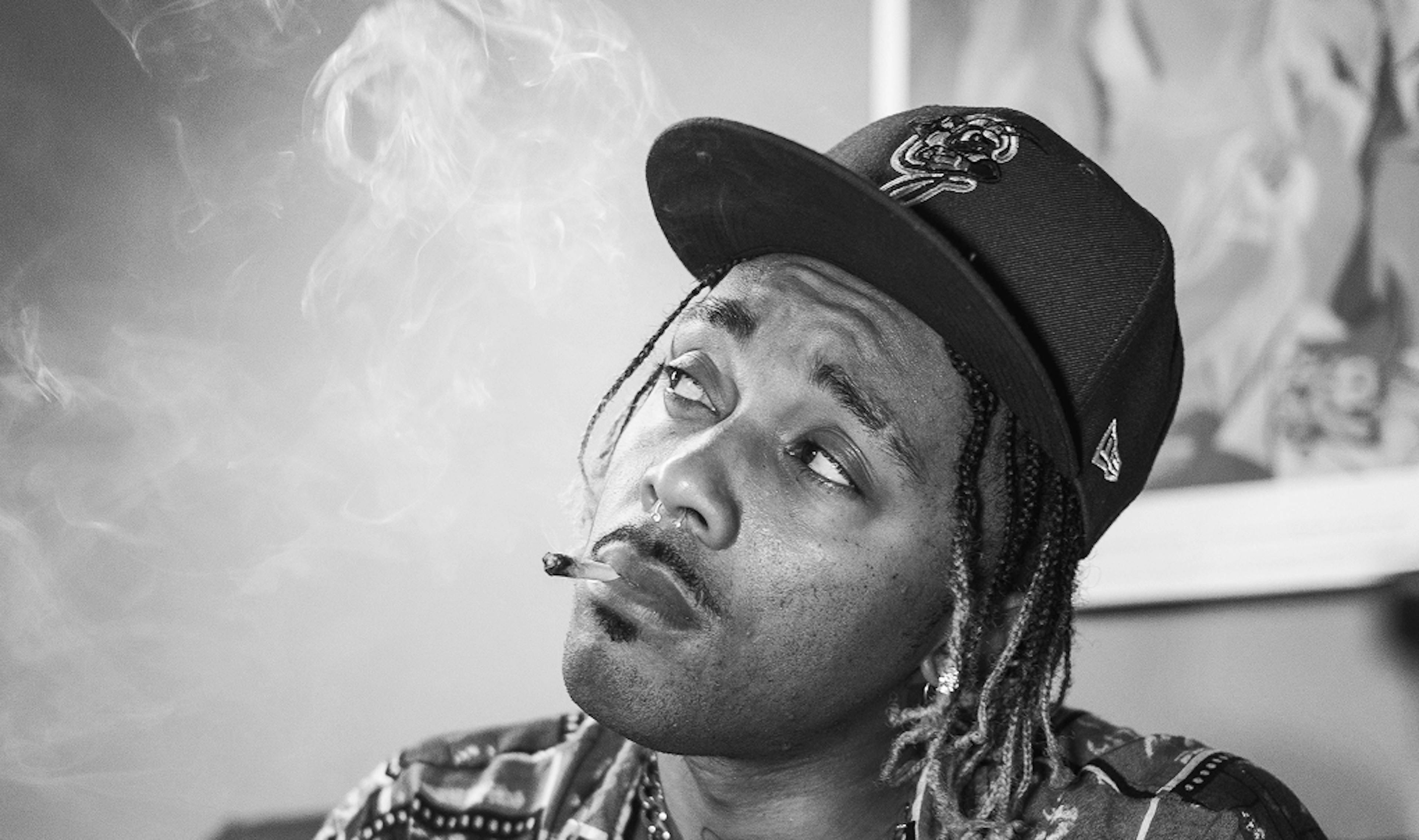 Black and white portrait of a man smoking a joint, wearing a patterned shirt and baseball cap, with smoke swirling around his face.