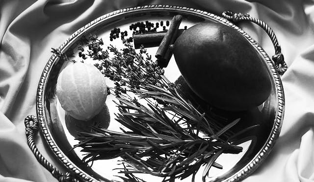 Close-up of a silver tray with natural ingredients including a lemon, dried lavender, cinnamon sticks, peppercorns, and a dark mango, arranged on a silky fabric backdrop.
