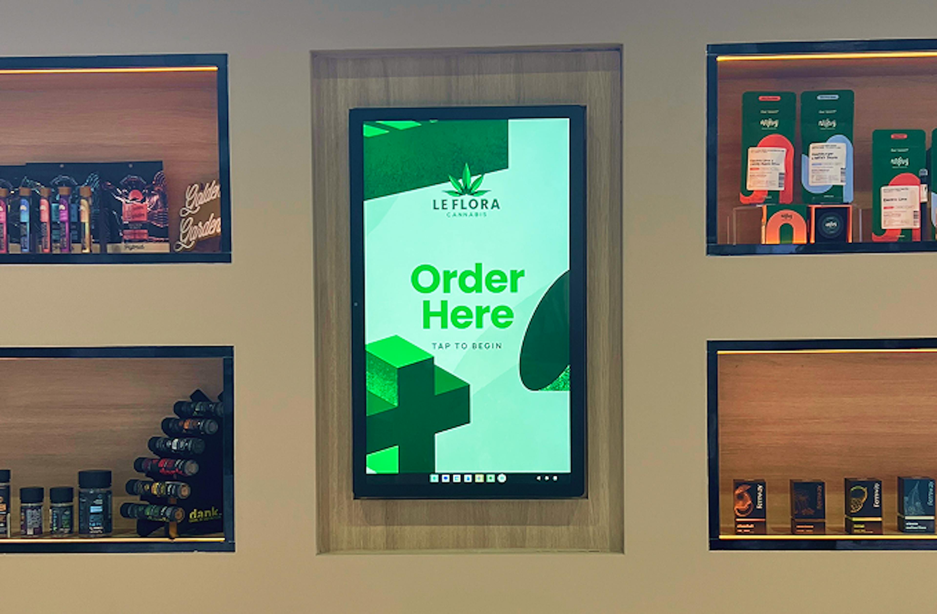 A touchscreen kiosk display inside a dispensary, framed with light wood and surrounded by product shelves. The screen shows a bright green interface with the Le Flora Cannabis logo and the text “Order Here – Tap to Begin.” Surrounding shelves display vape cartridges, pre-rolls, and branded cannabis products.