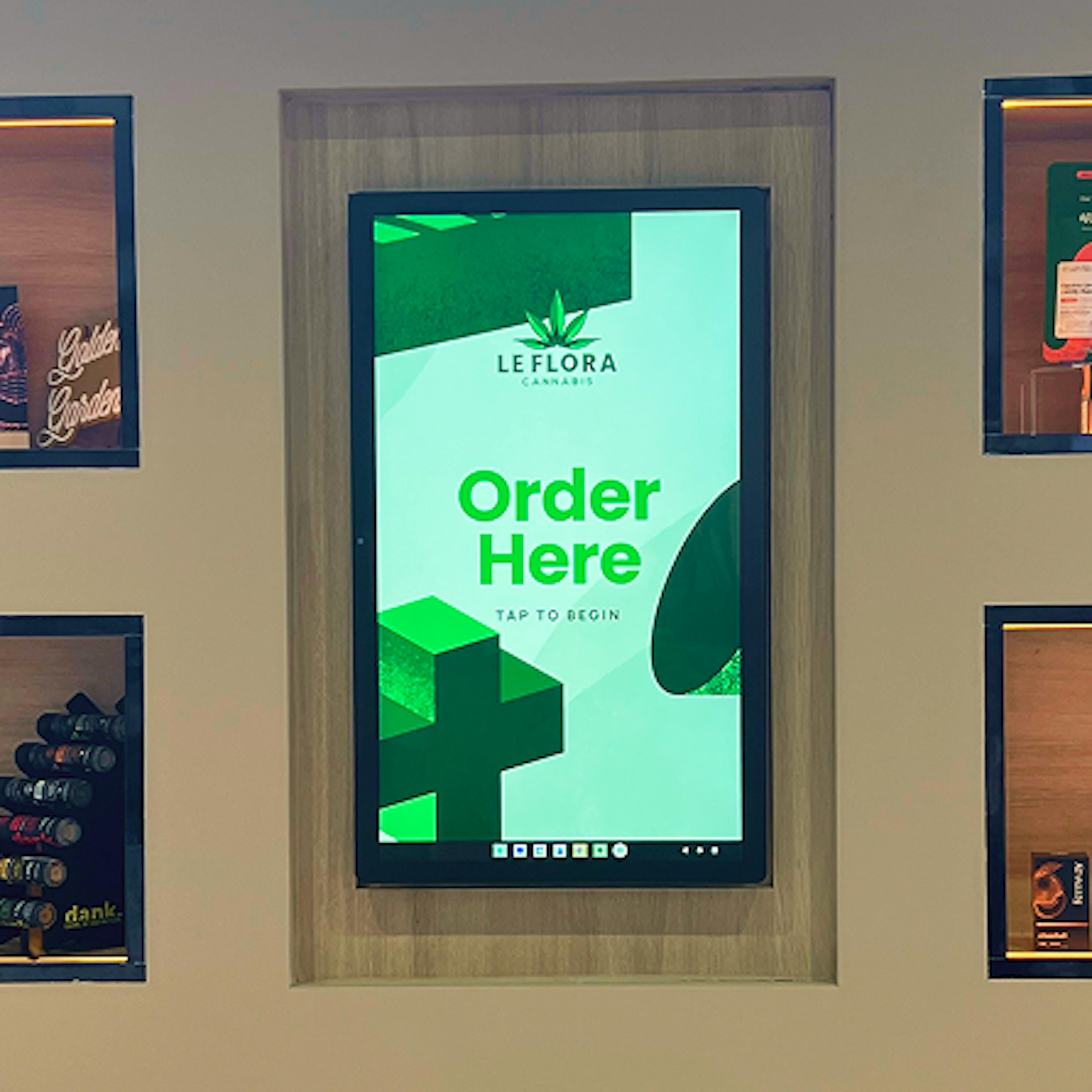 A touchscreen kiosk display inside a dispensary, framed with light wood and surrounded by product shelves. The screen shows a bright green interface with the Le Flora Cannabis logo and the text “Order Here – Tap to Begin.” Surrounding shelves display vape cartridges, pre-rolls, and branded cannabis products.
