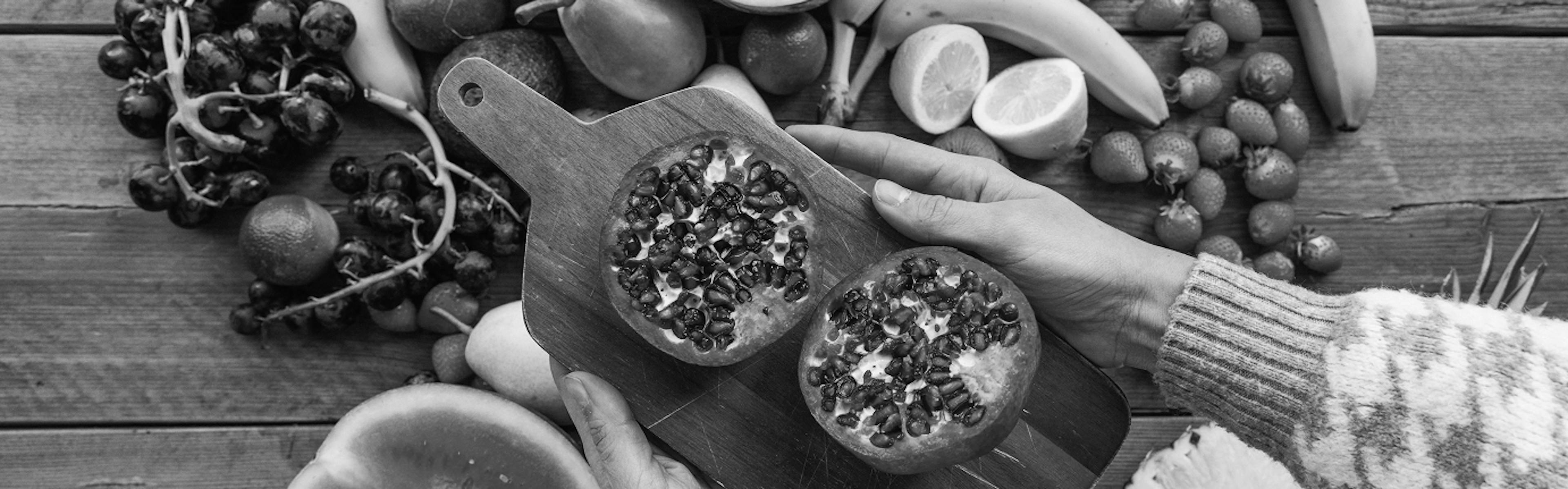 Hands holding a wooden board with halved pomegranates over a rustic table filled with bananas, watermelon, grapes, and other fruits.