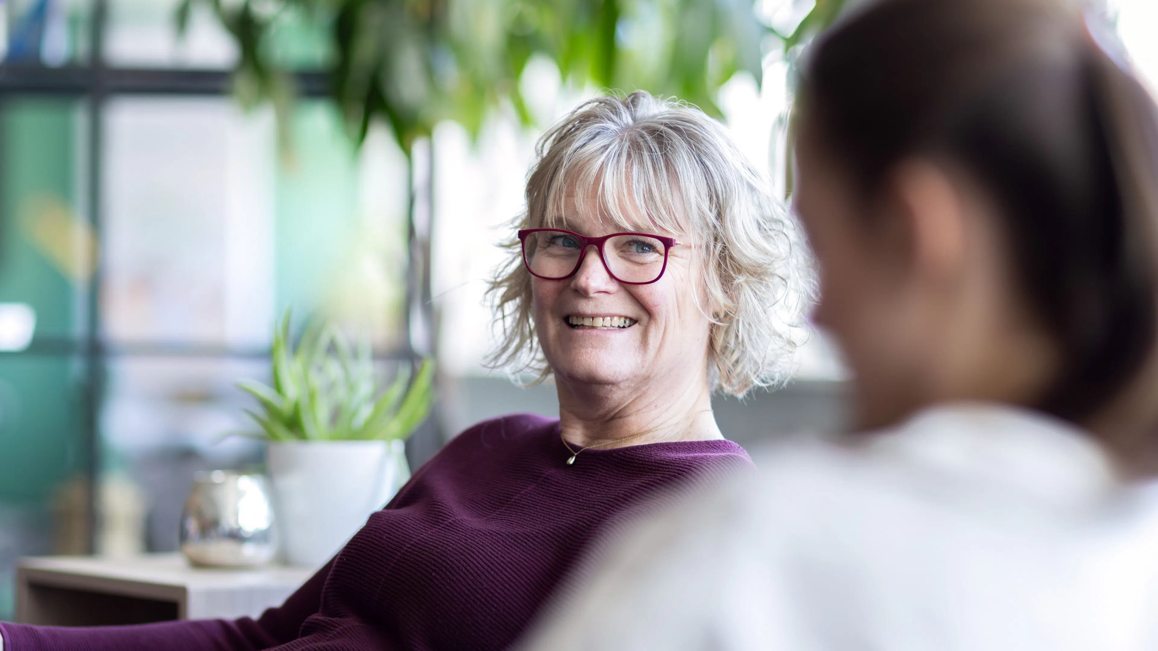 Kvinna sitter i en soffa har lila tröja och glasögon.
