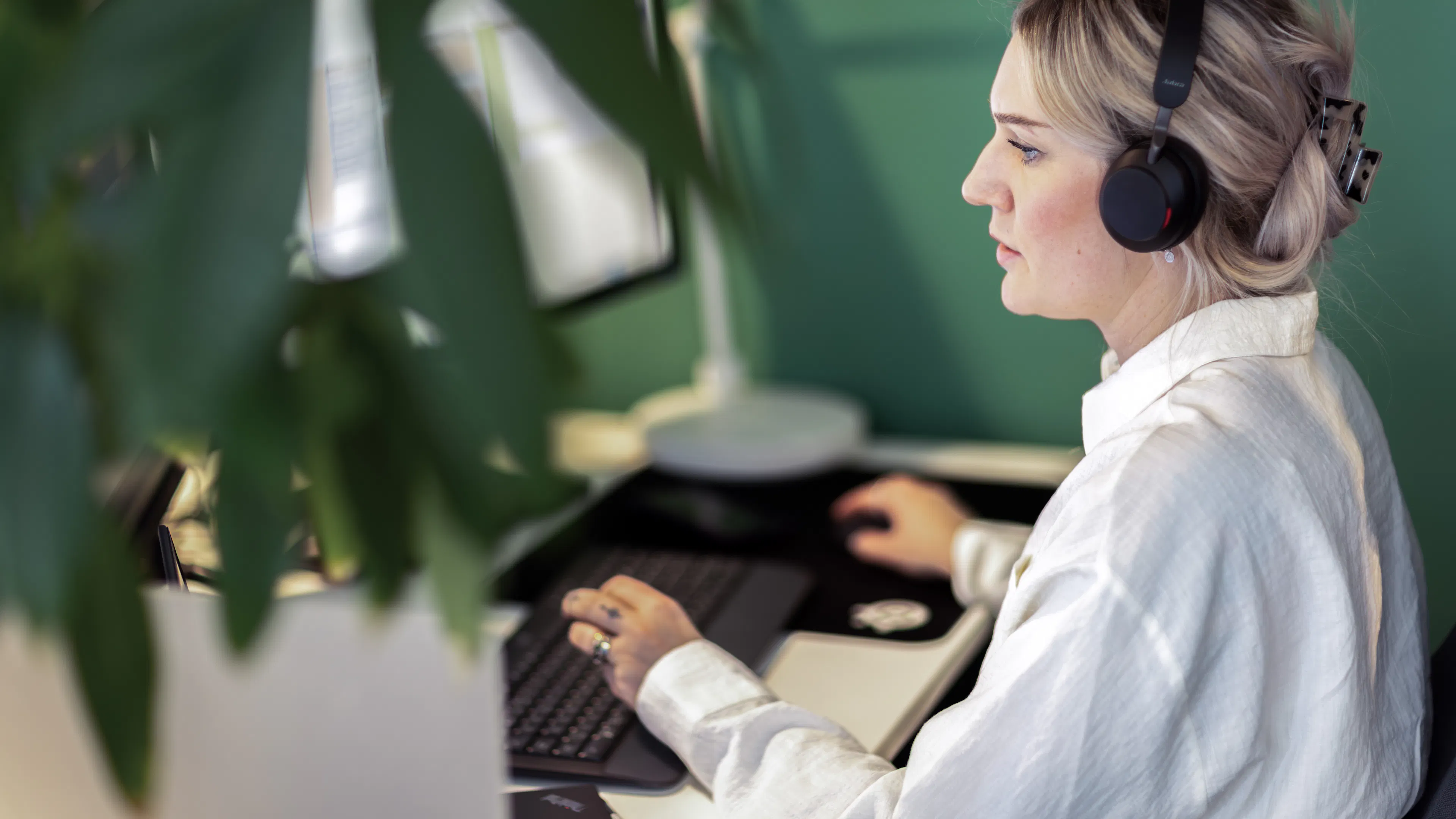 Kvinna i vit skjorta och headset sitter framför en dator.