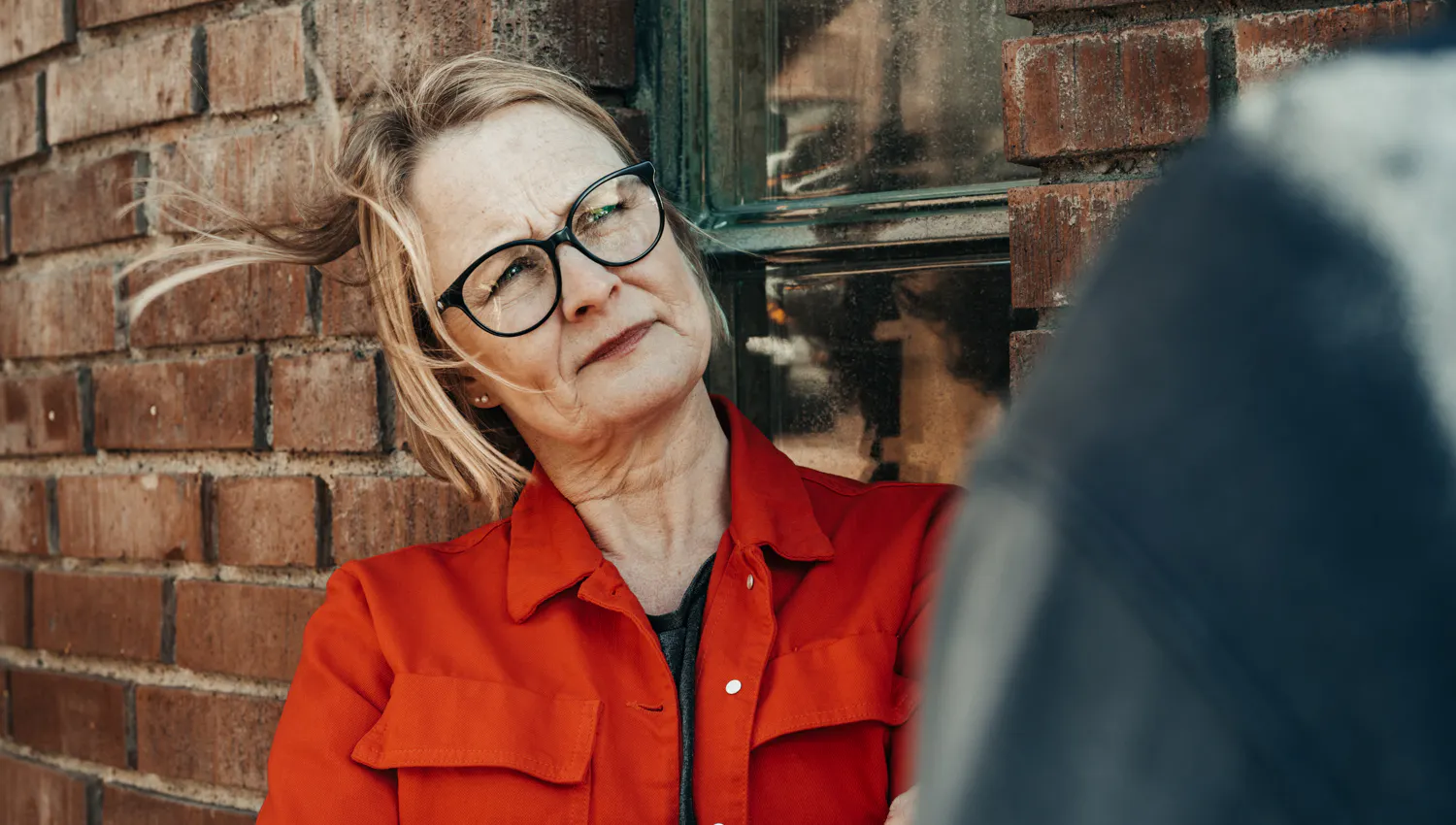 Kvinna i röd jacka med glasögon och vind i håret.