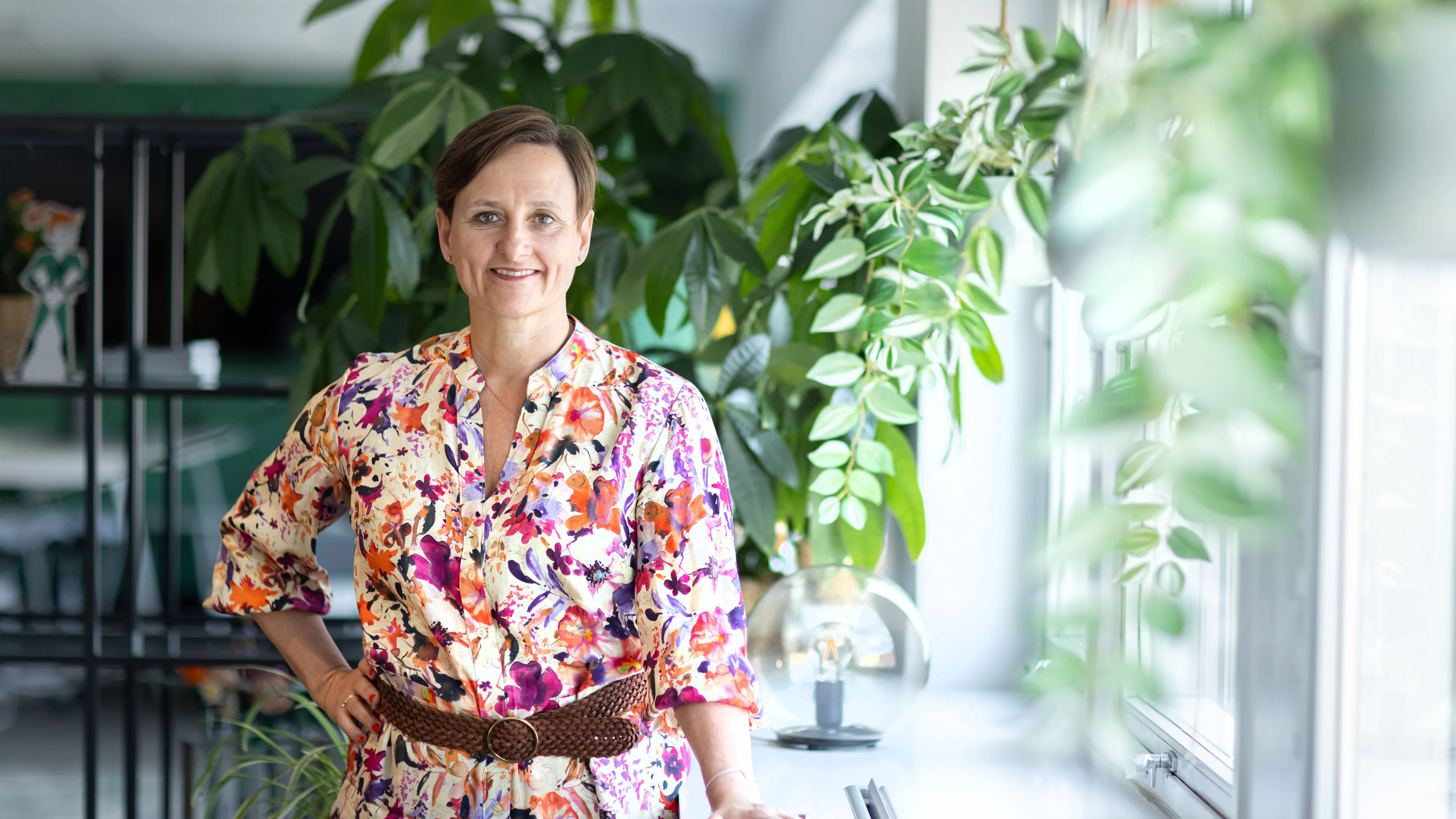 Kvinna med blommig klänning som står framför gröna växter.