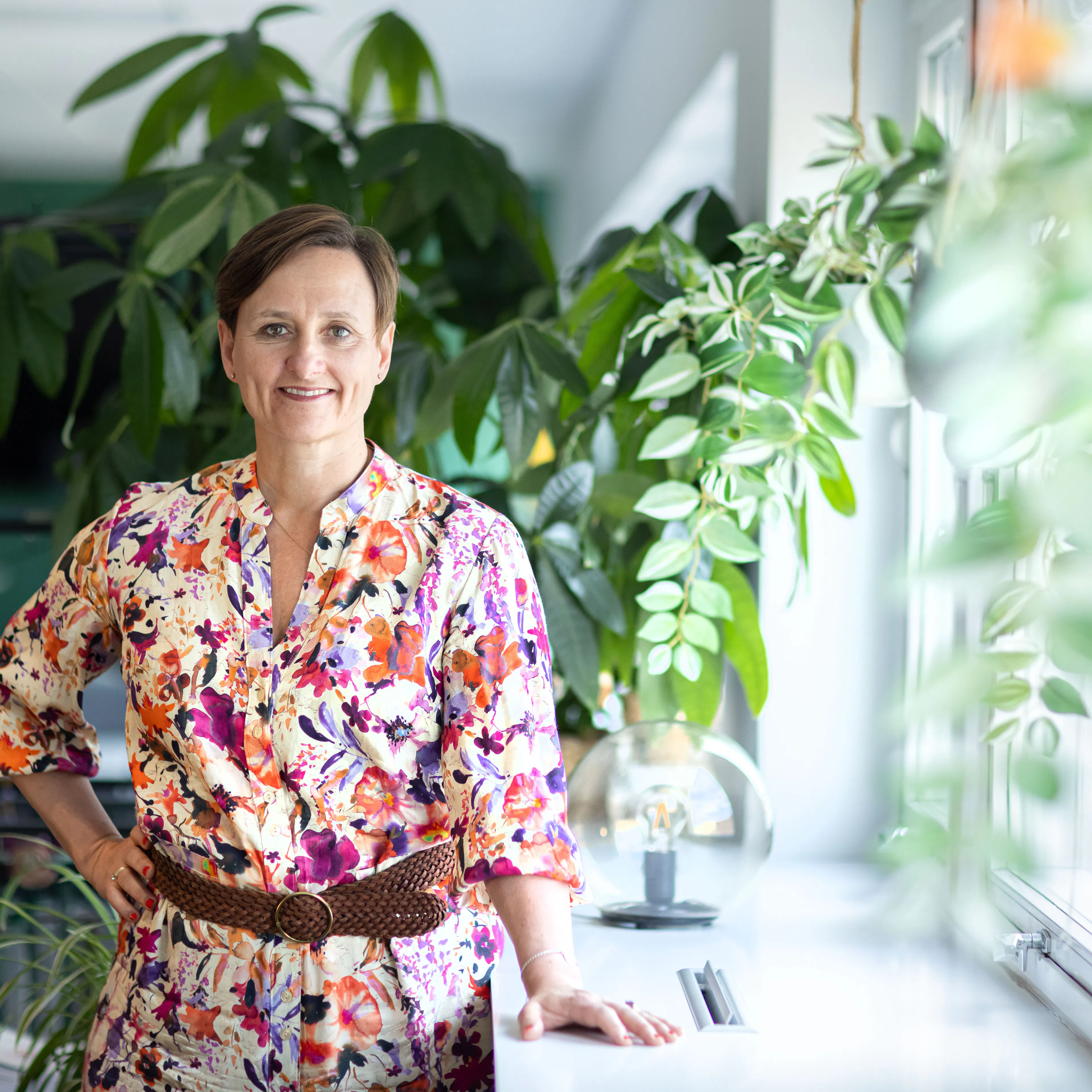 Kvinna med blommig klänning som står framför gröna växter.